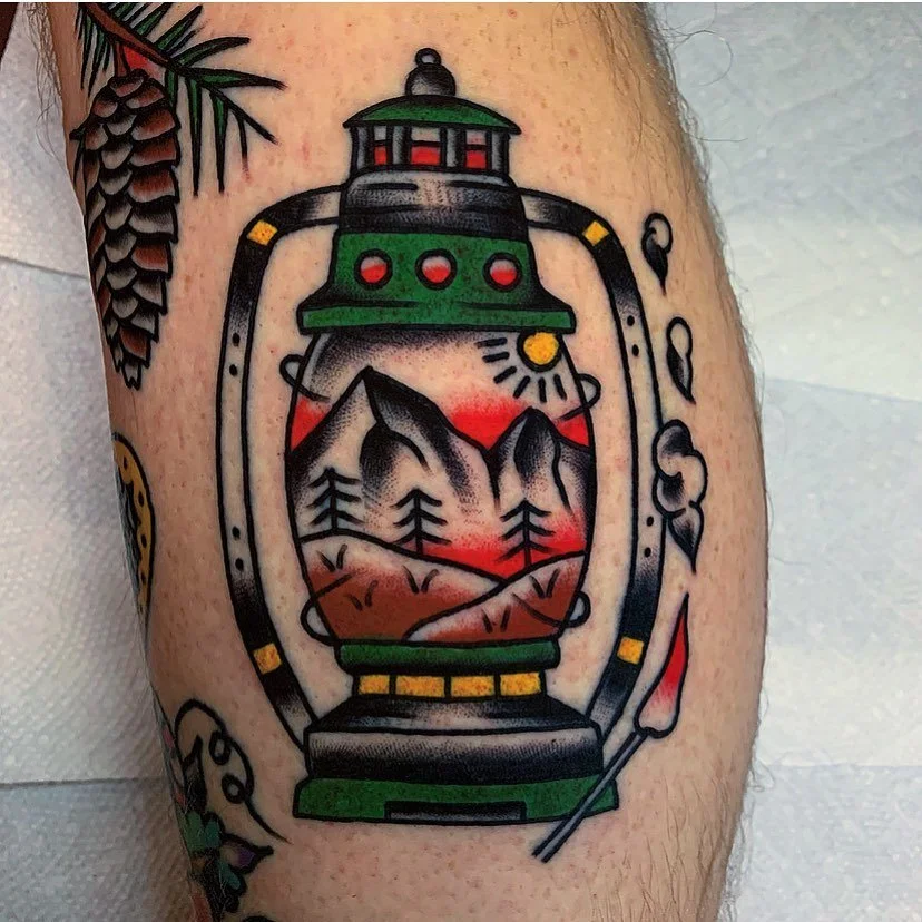 Tattoo of a lantern with a landscape scene including mountains, trees, the sun, and red sky, situated on a person's arm.
