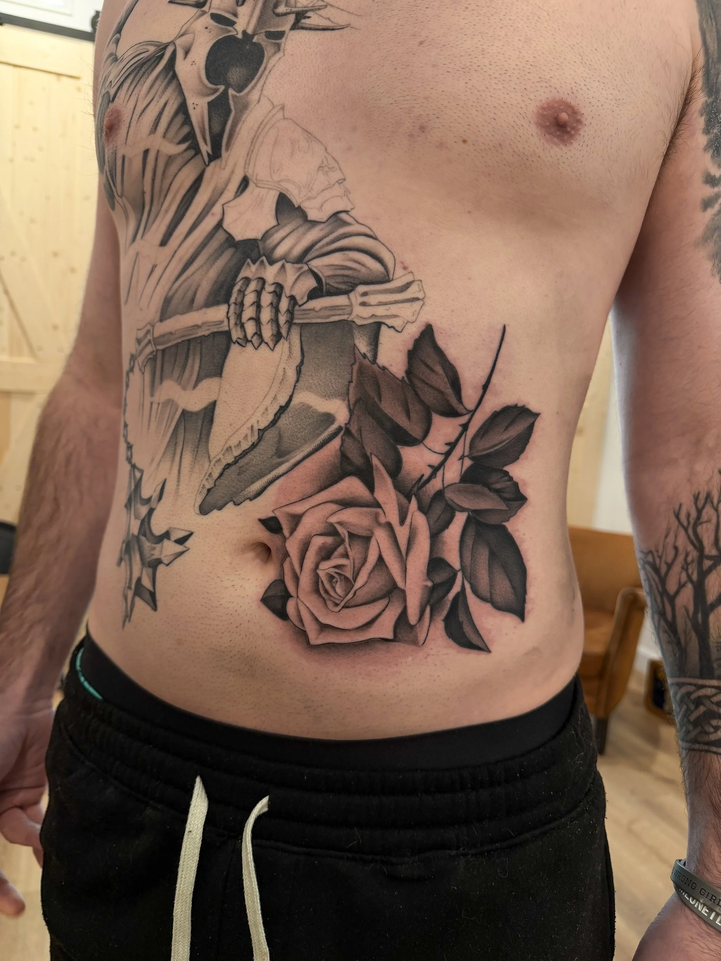 A person's side torso with a black and gray tattoo of roses, leaves, and a skeleton bird wearing a hooded cloak.