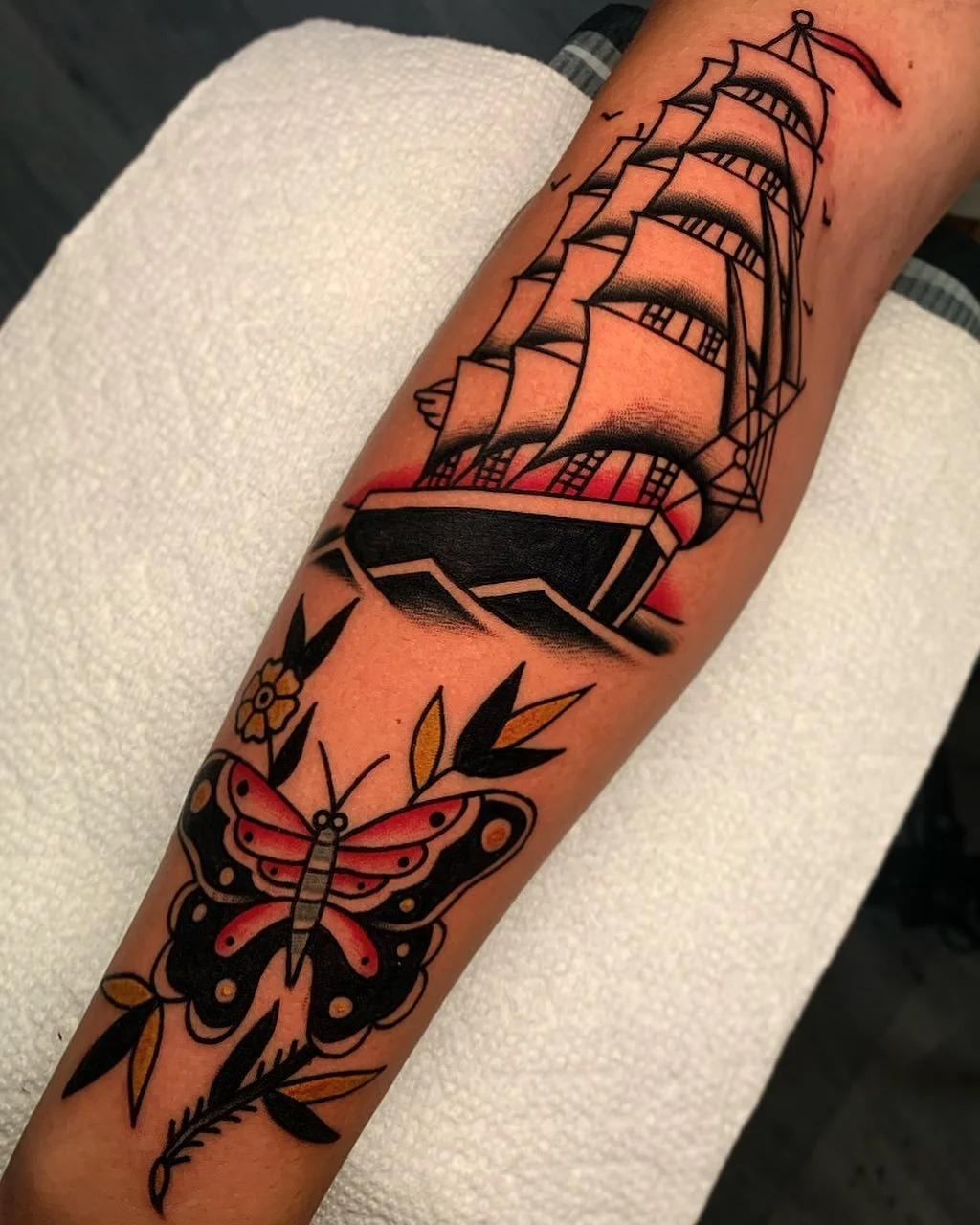 Tattoo of a sailing ship with full sails, accompanied by a pink and black butterfly and yellow leaves.