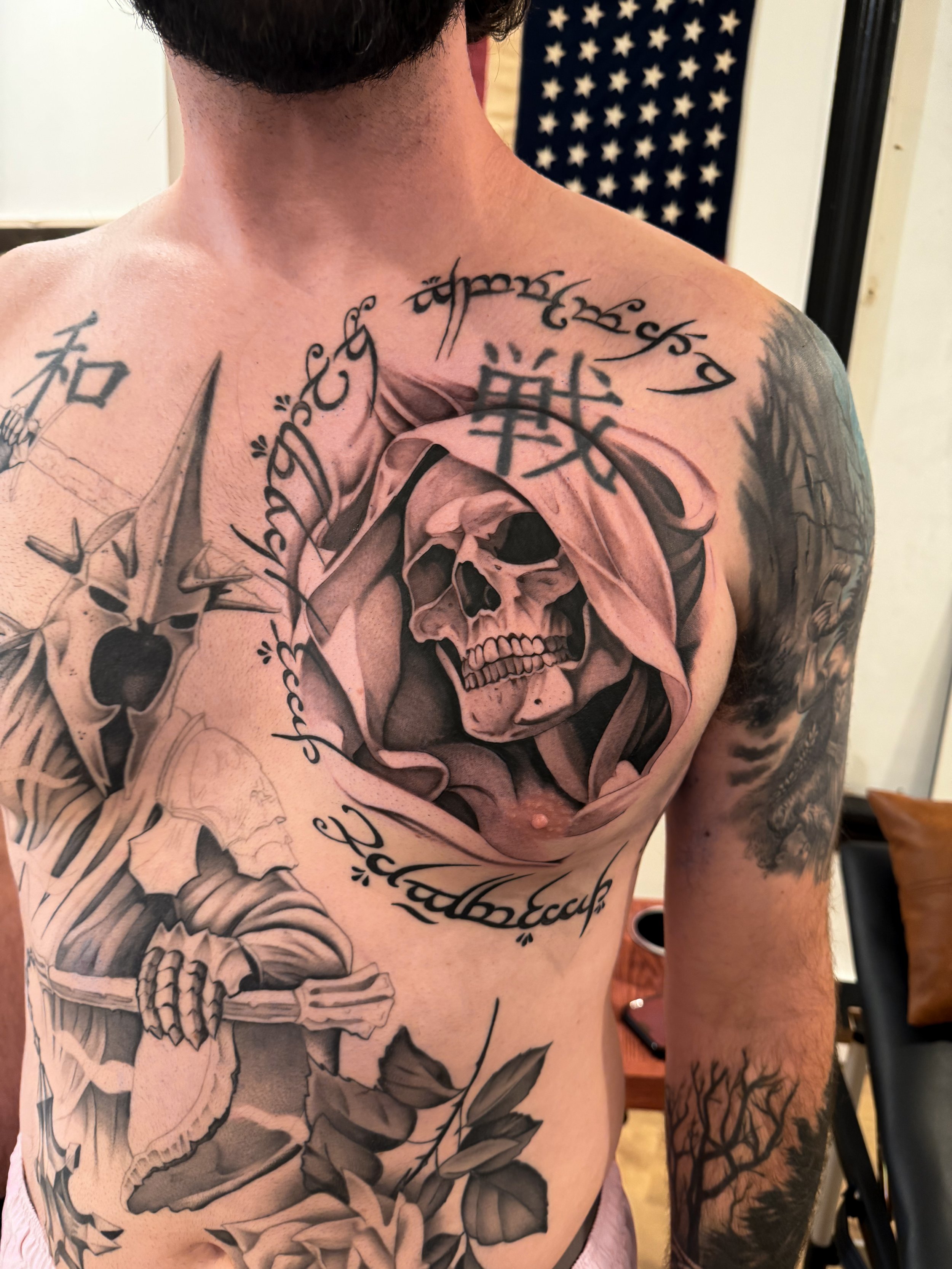 A person's upper chest and arm with multiple tattoos, including a skull, Japanese characters, and decorative elements. An American flag is partially visible in the background.