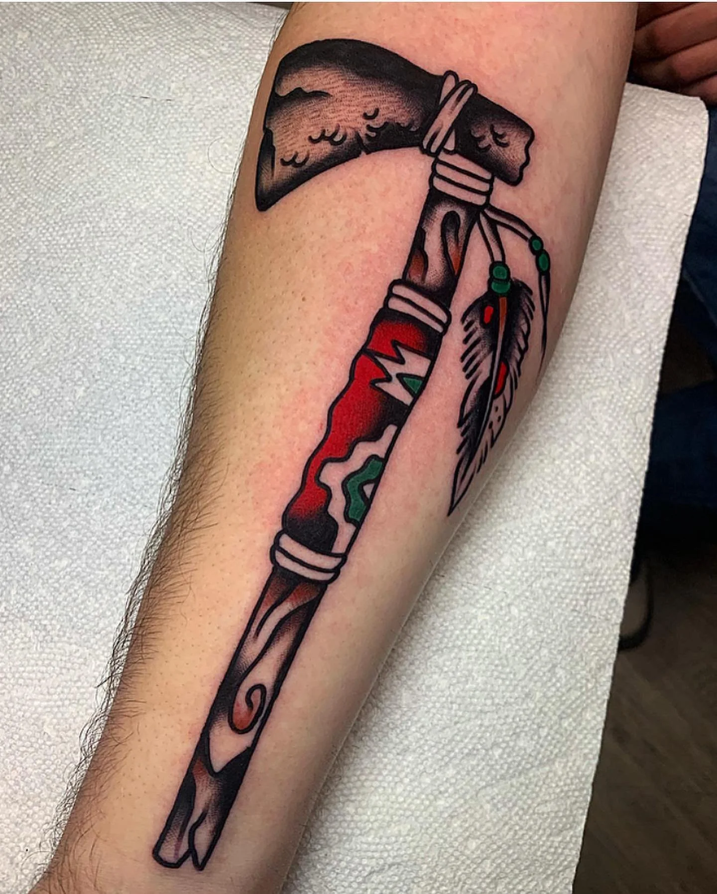 Tattoo of a knife with a rainbow-colored lightning bolt design on the blade, wrapped with a feather and beads.