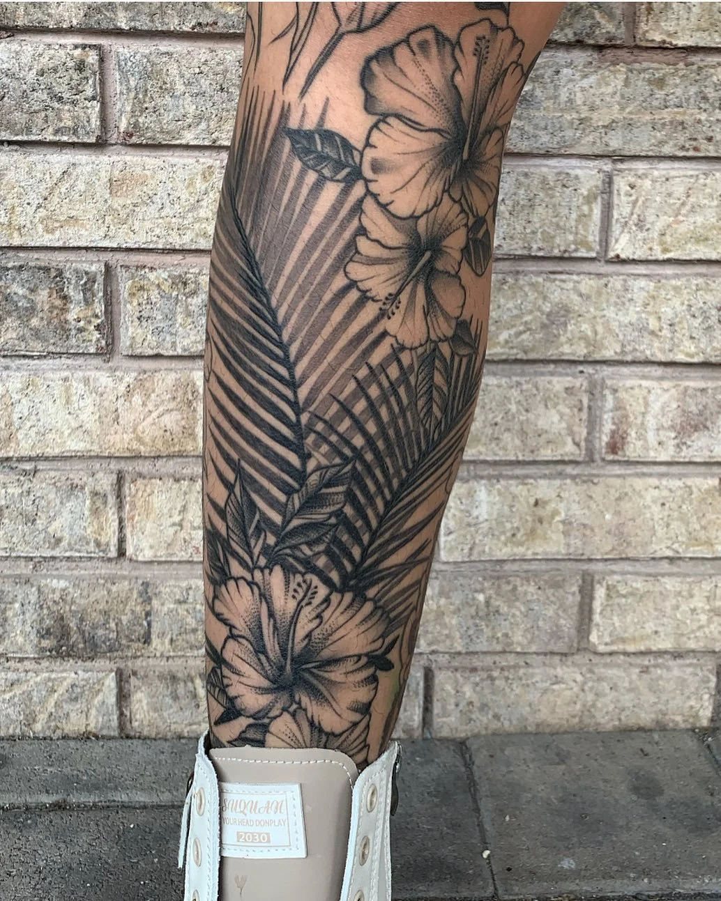Black and gray floral tattoo on a person's leg featuring large hibiscus flowers and palm leaves.