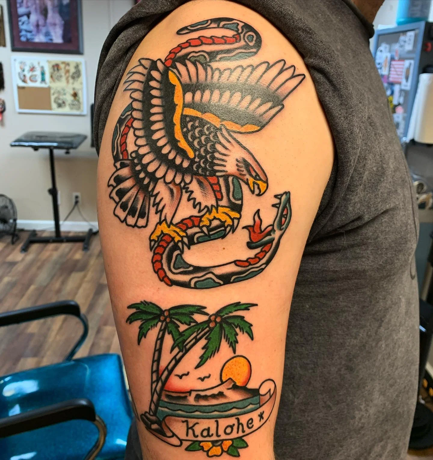 Tattoo of an eagle with outstretched wings, a snake in its claws, a tropical palm tree, the sun setting over the ocean, and the word ‘Kolohe’ on a scroll at the bottom.