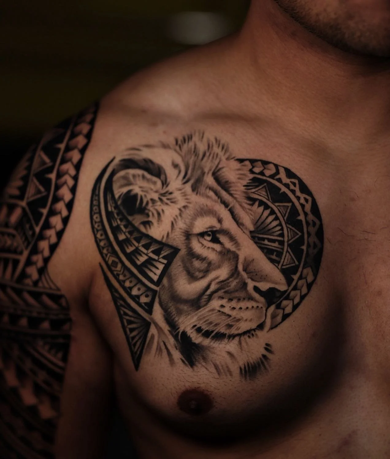 Close-up of a man's chest with a black tribal-style tattoo featuring a realistic lion's face surrounded by geometric patterns.
