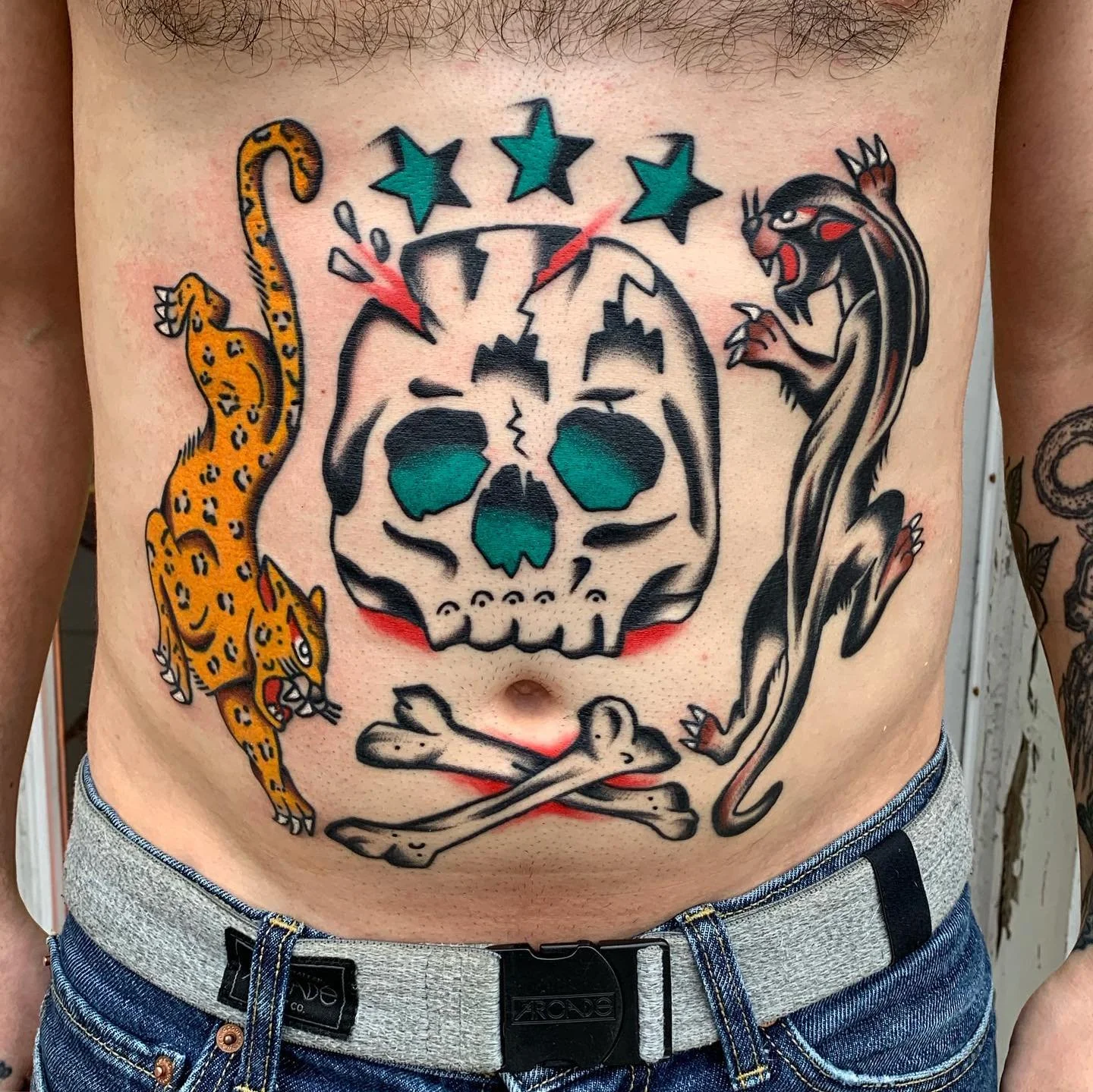 Tattoo of a skull with turquoise eye sockets, surrounded by two jumping jaguars, one on each side, with four green stars above the skull.