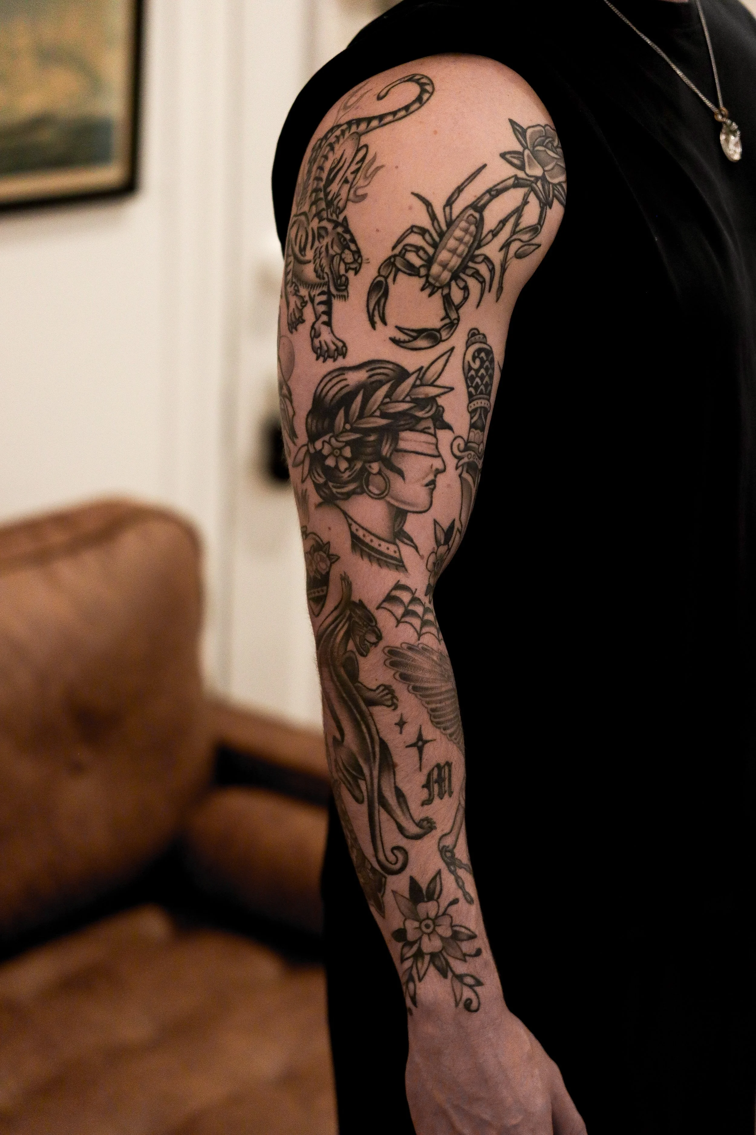 Close-up of a person's arm with various black tattoo designs, including a tiger, a scorpion, a woman's profile, and flowers.
