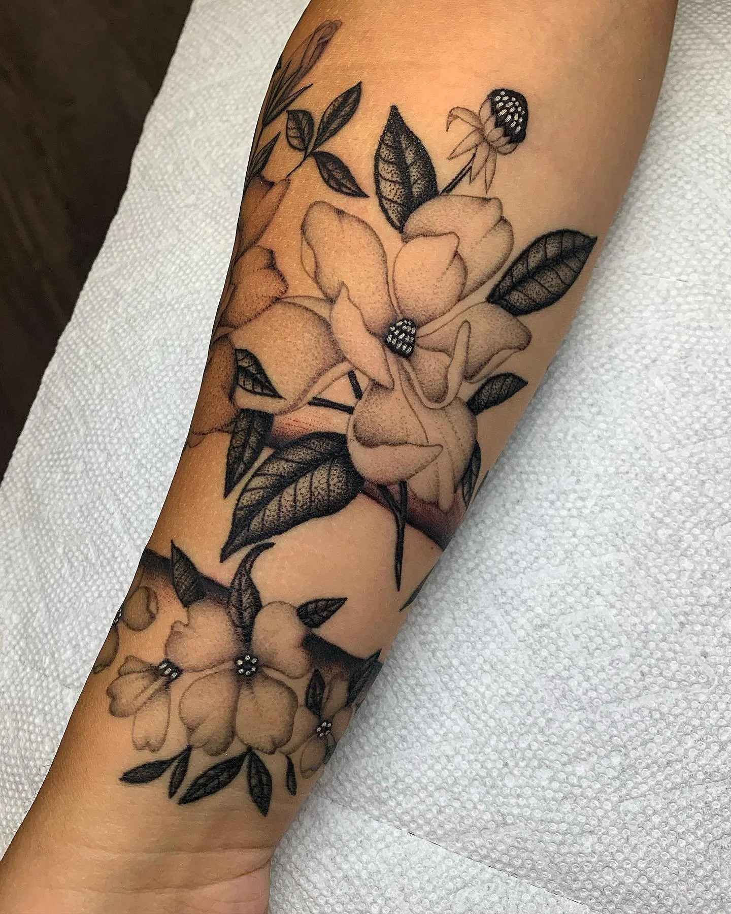 Black and gray floral tattoo on a person's arm, featuring various flowers and leaves with intricate shading.