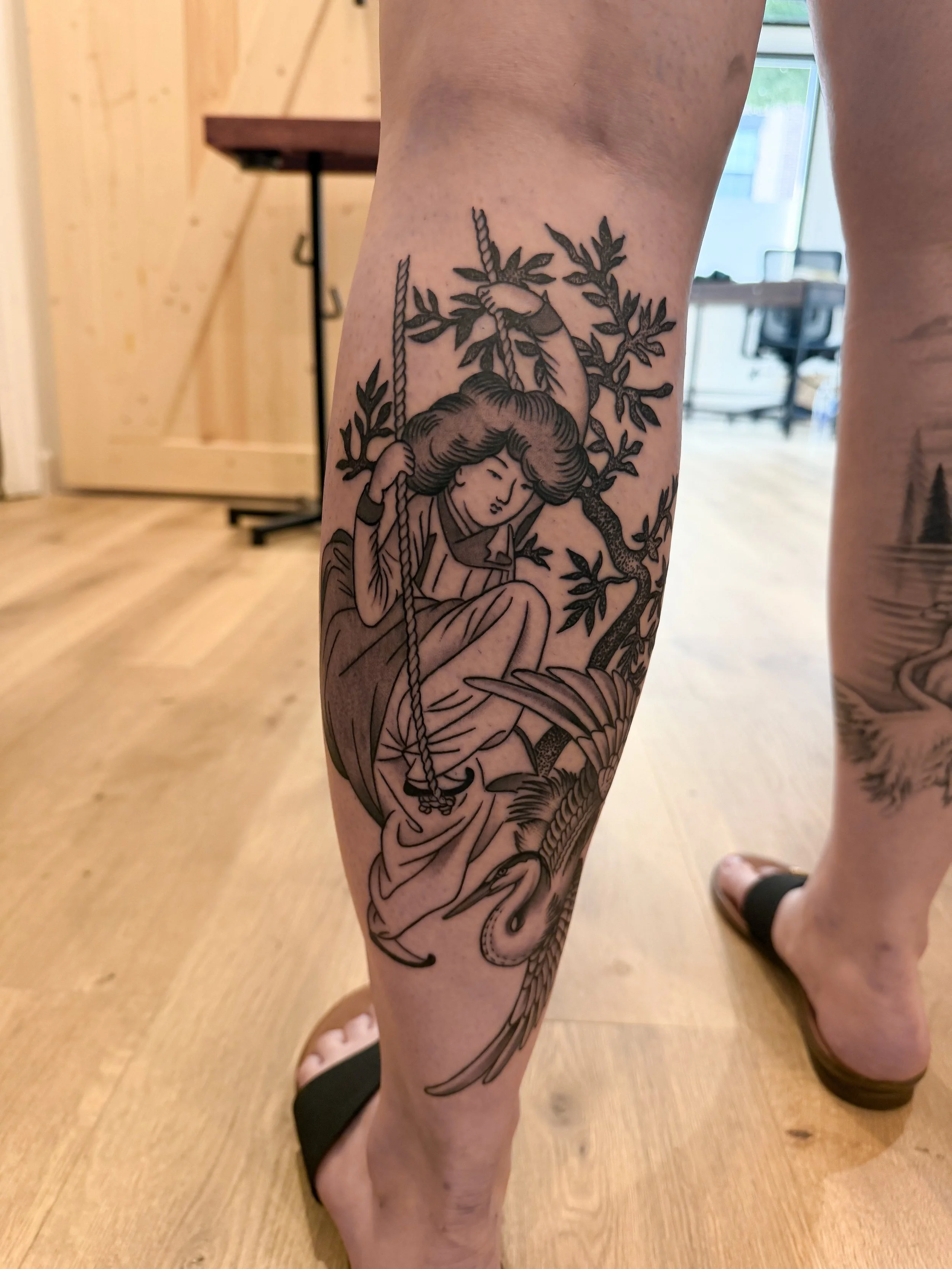 Tattoo of a woman with long hair sitting under a tree with rope swing, on a person's leg.