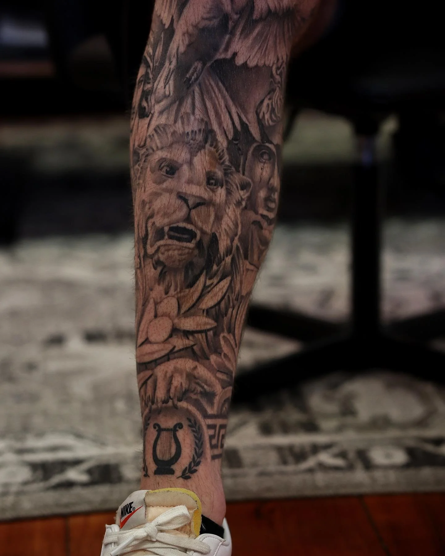 Close-up of a person's lower leg and foot, showing a detailed tattoo of a lion's face and other designs, wearing a white Nike sneaker.