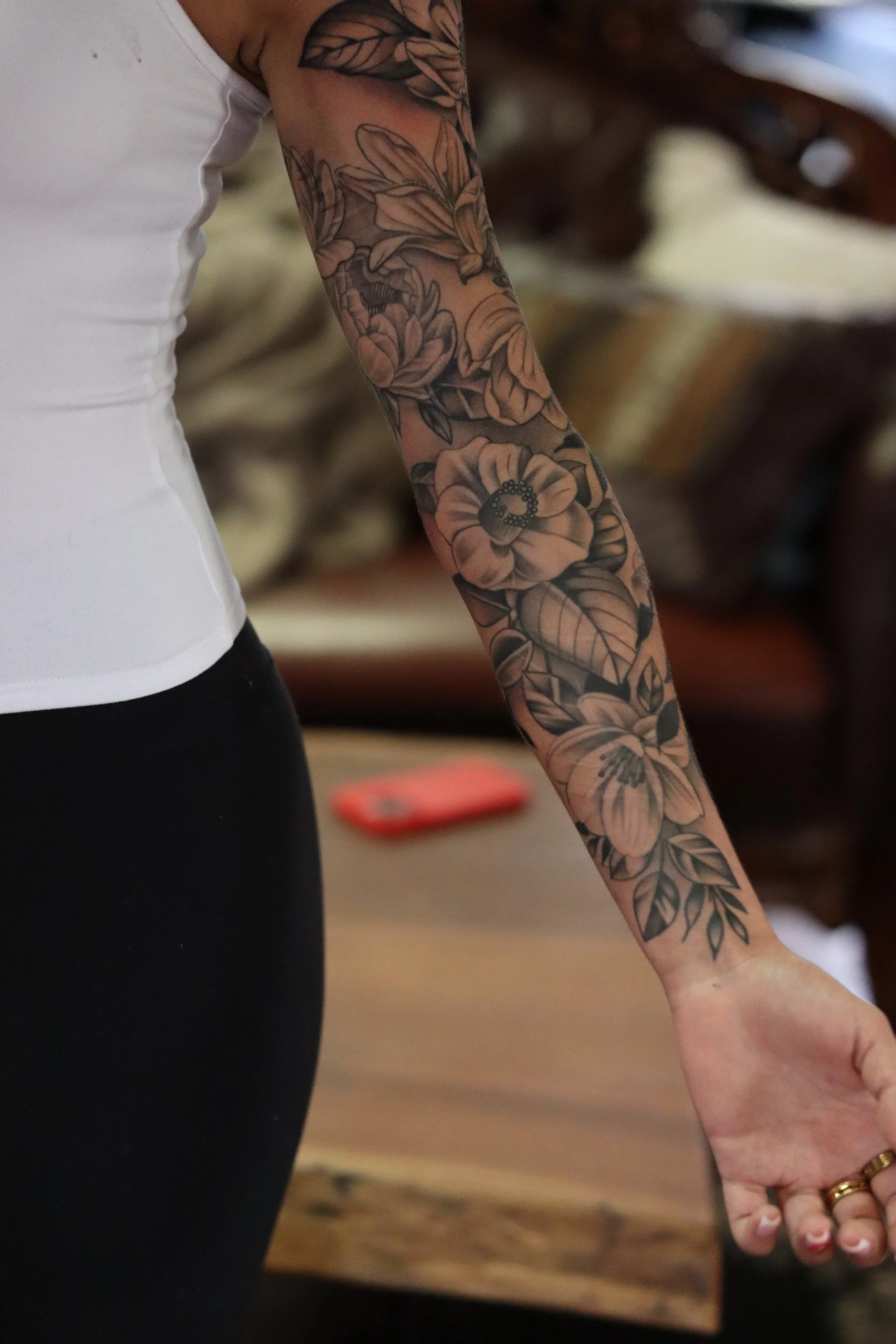 A person's arm adorned with a detailed black and gray floral tattoo, extending from the shoulder to the wrist, with the person wearing a white sleeveless top.