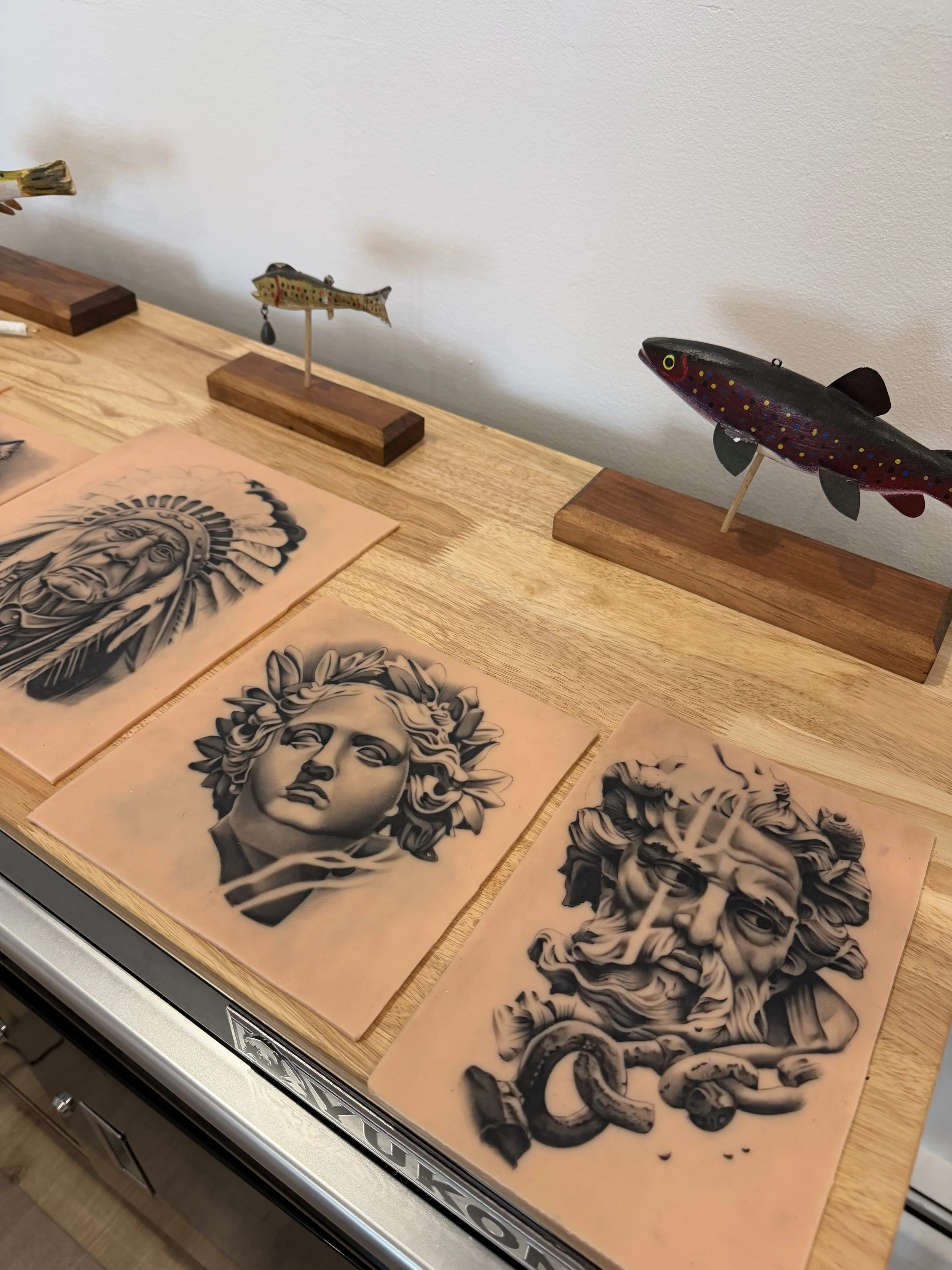 Wooden table displaying four black and white tattoo stencil sheets with realistic portrait and floral designs.
