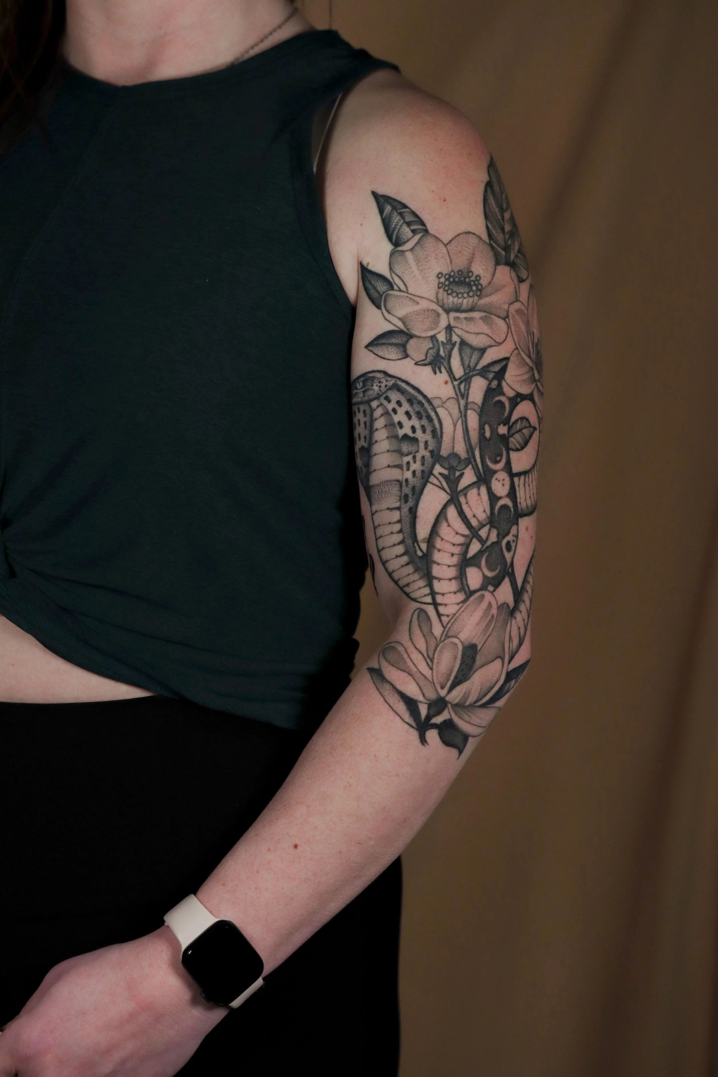 A woman wearing a black sleeveless top with a floral tattoo on her upper arm and a smartwatch on her wrist.