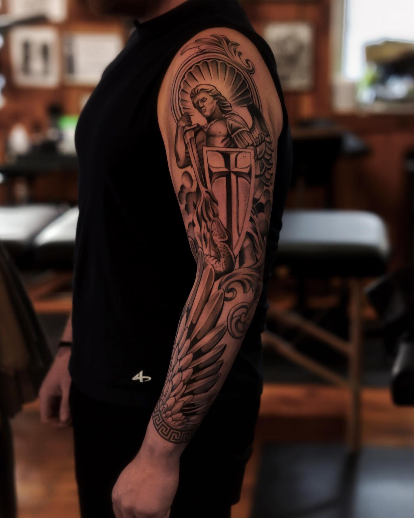 Black and gray tattoo of a female warrior holding a sword and shield, with a halo-like radiance behind her and dragon scales on her arm, on a person's upper arm.