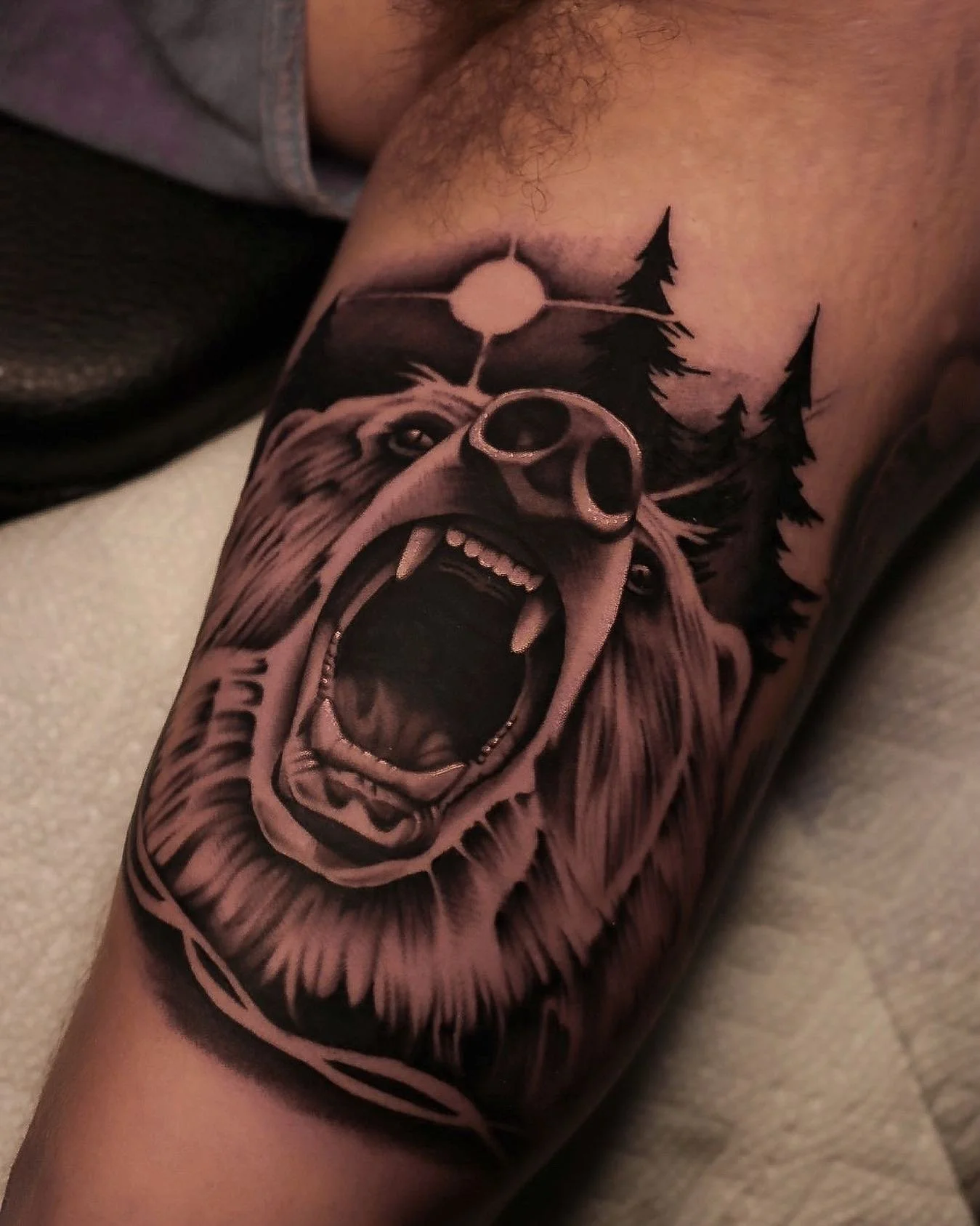 Tattoo of a roaring bear with trees and a moon in the background on a person's arm.