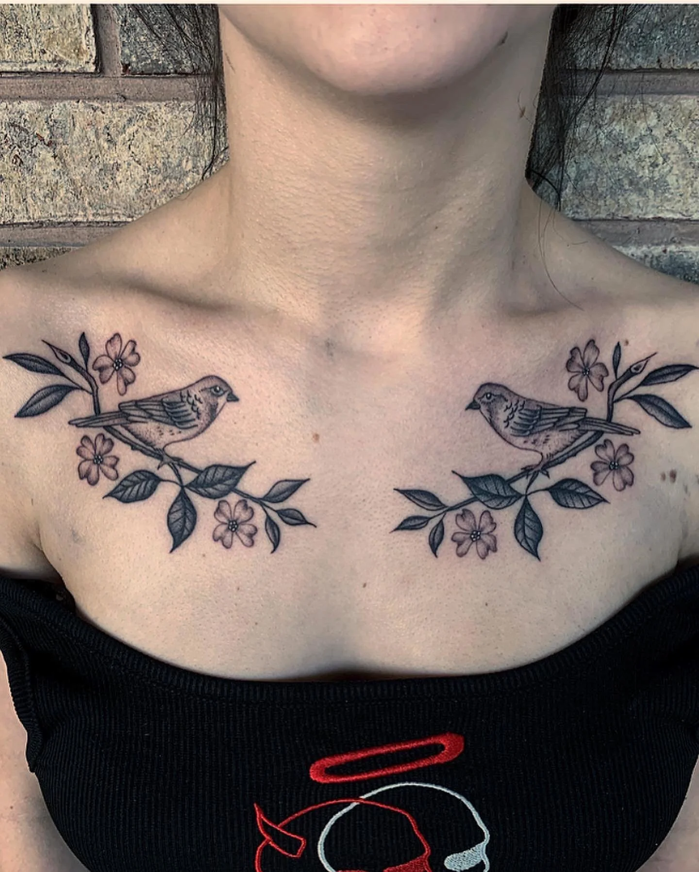 Black tattoo of two birds sitting on branches with leaves and flowers, placed across the collarbone area of a person with light skin.
