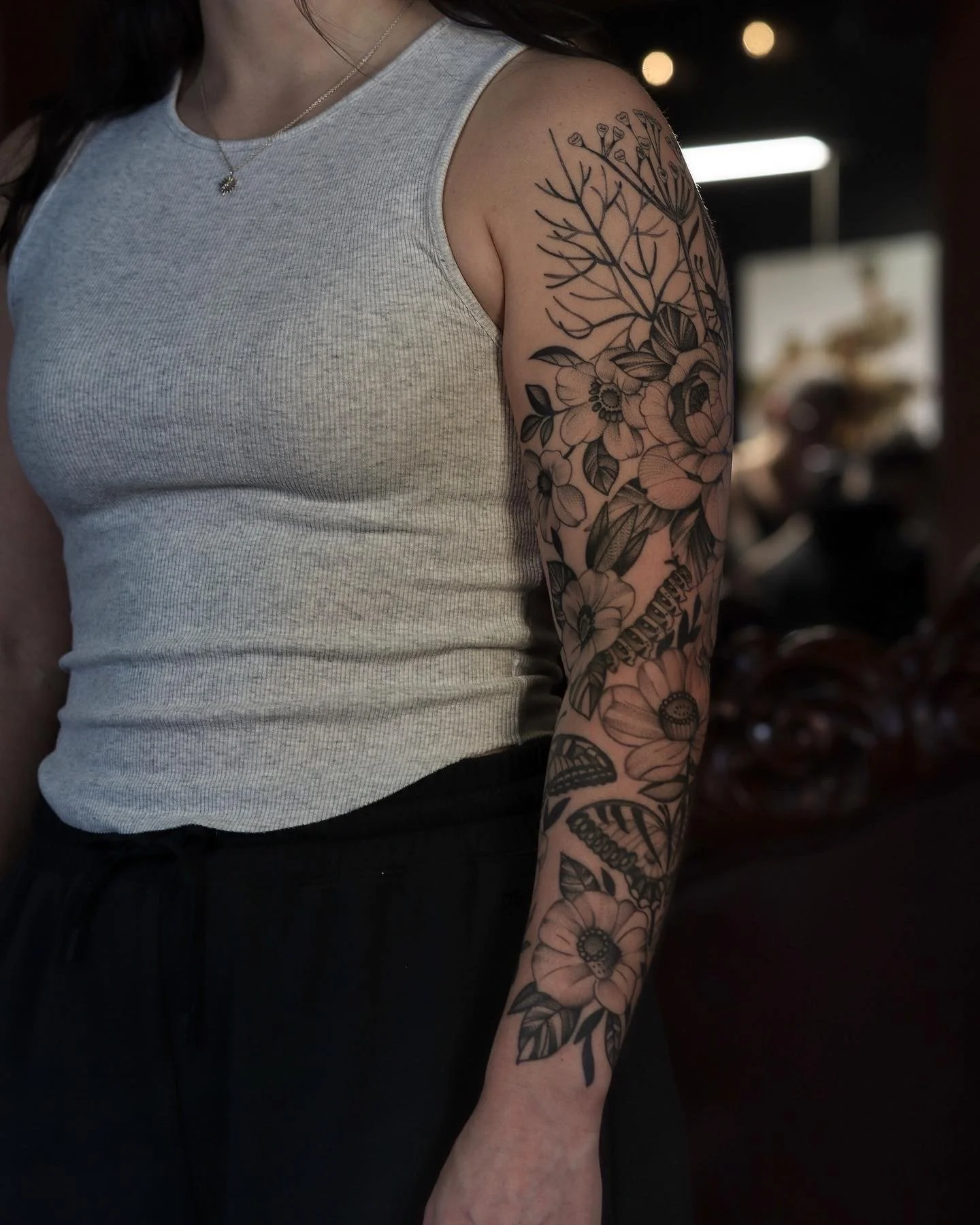 A person wearing a sleeveless gray top and black pants with a detailed black floral tattoo sleeve on their left arm, standing indoors with blurred background.