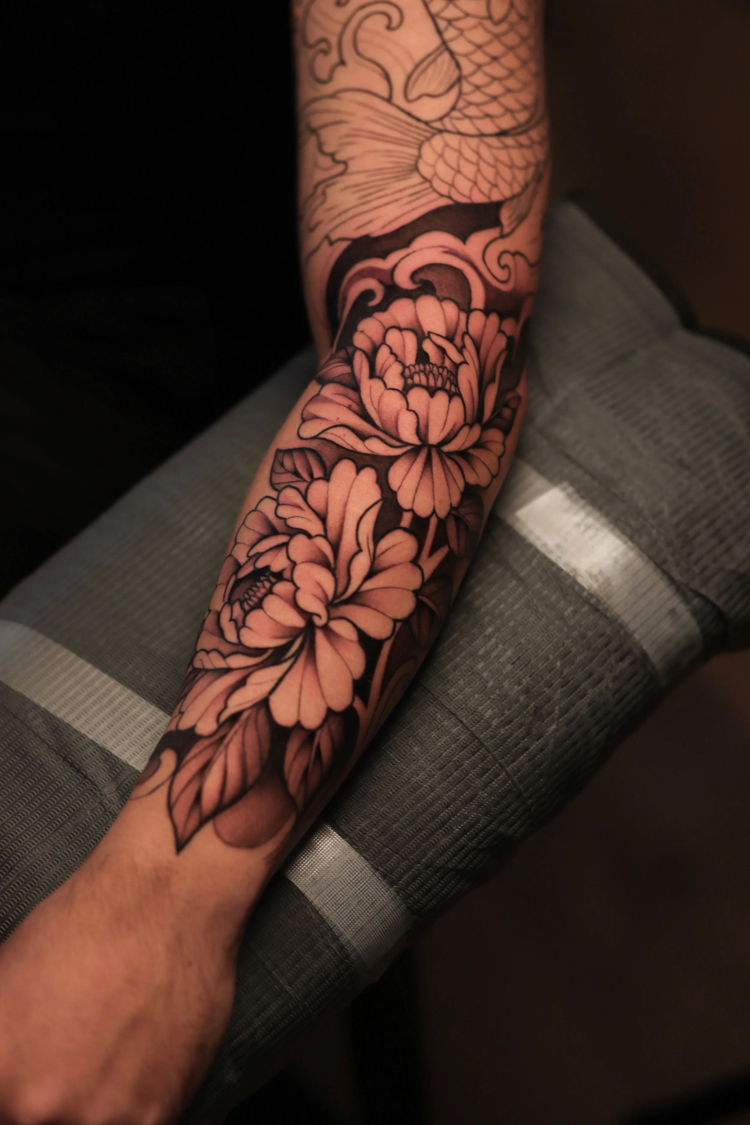 Tattoo sleeve featuring a koi fish swimming above large blooming peonies with detailed leaves, all in black ink.