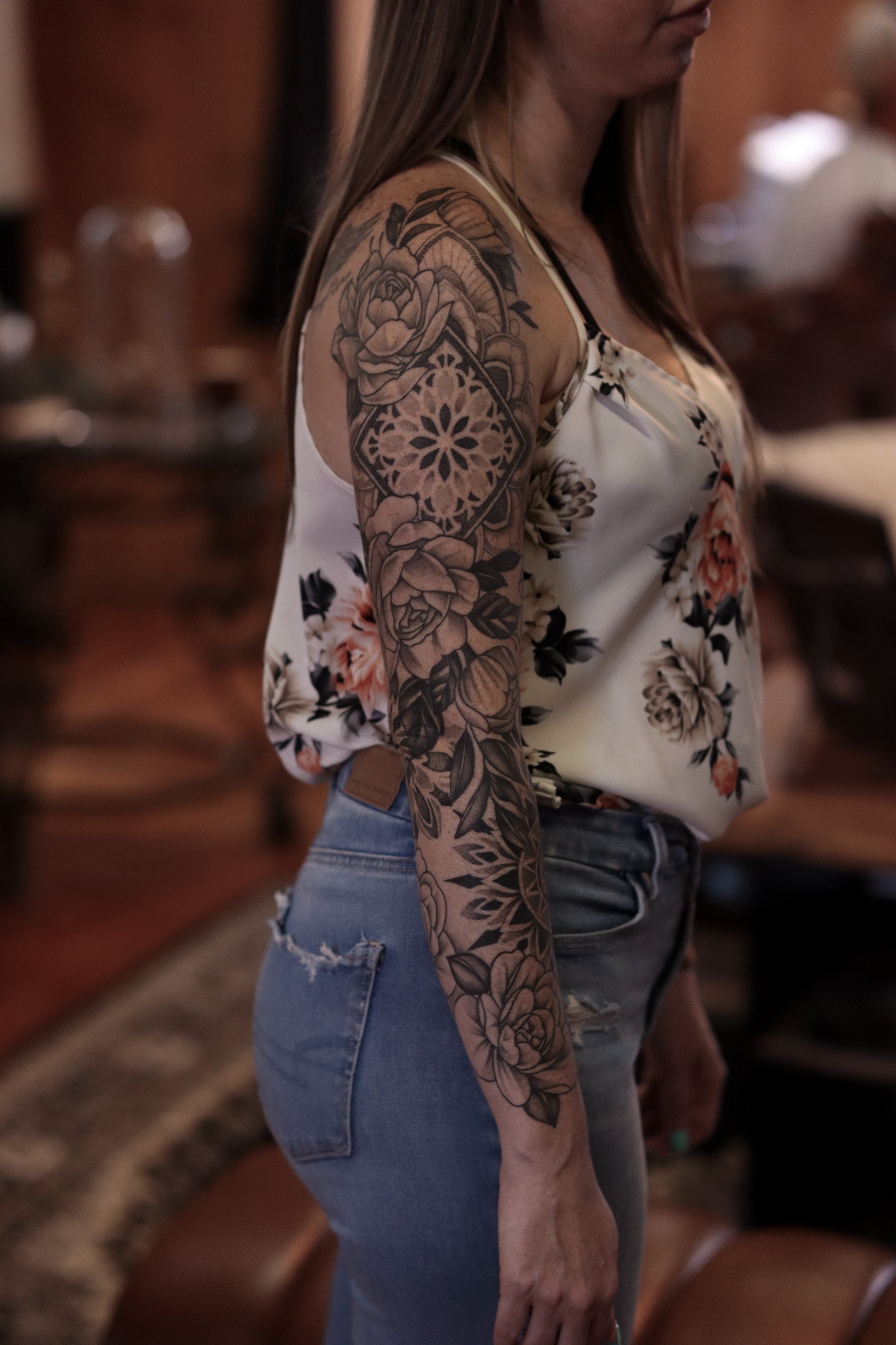 A woman with long brown hair is shown in profile view, wearing a floral tank top and light blue ripped jeans. She has a large floral tattoo on her upper arm and forearm.