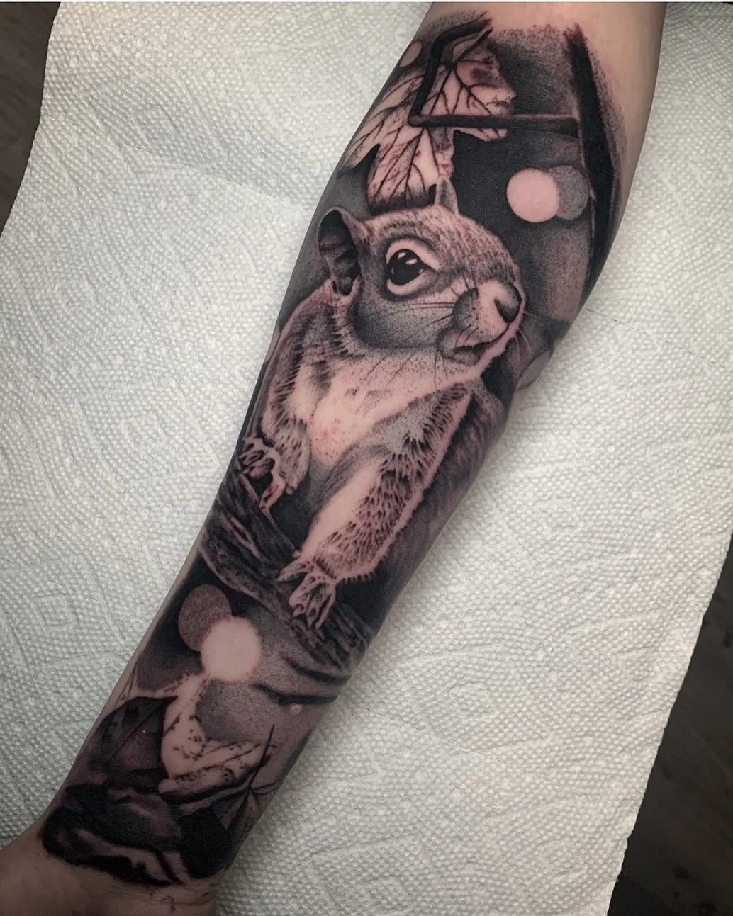 Black and gray tattoo of an owl and a small animal, possibly a rodent, on a person's arm. The owl appears to be sitting on a branch with a moon and a textured background.