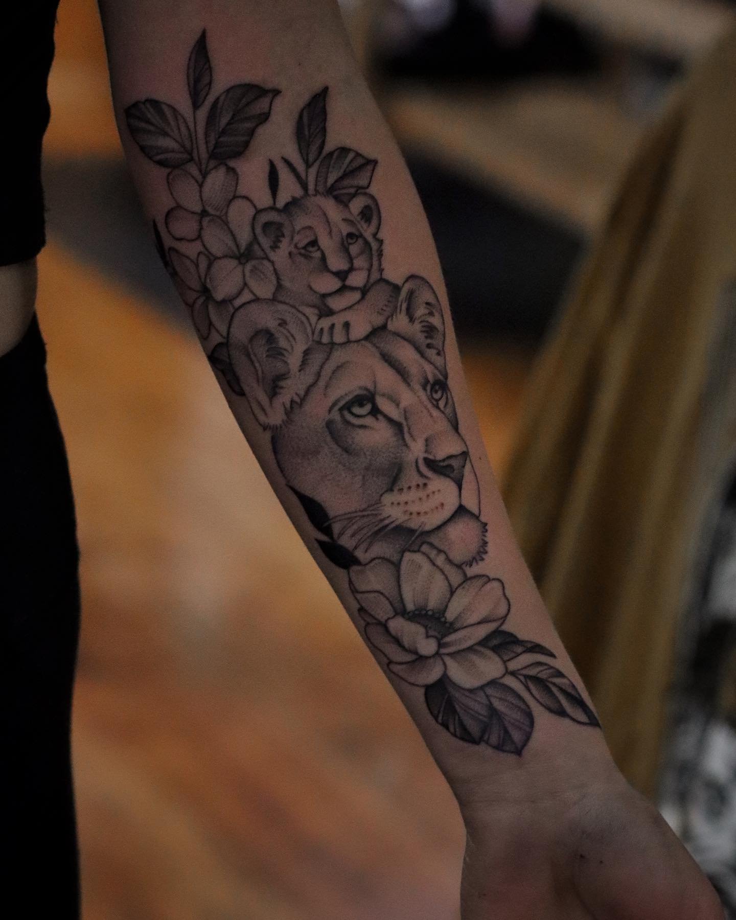 Black and gray tattoo of a lion's face, surrounded by flowers and leaves, on a person's arm.