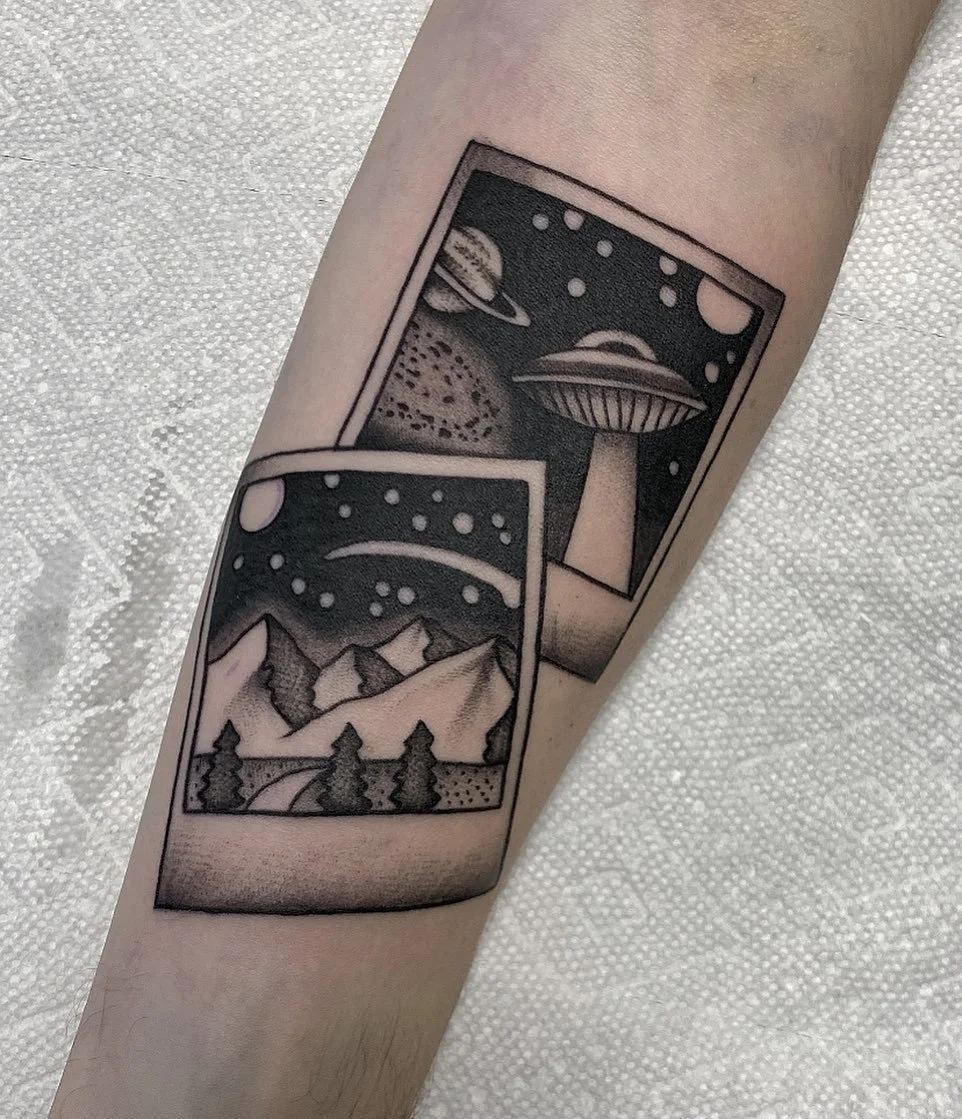 Black and gray tattoo of two rectangular panels, one showing a night sky with planets and stars, and the other showing mountains, trees, and a starry sky.