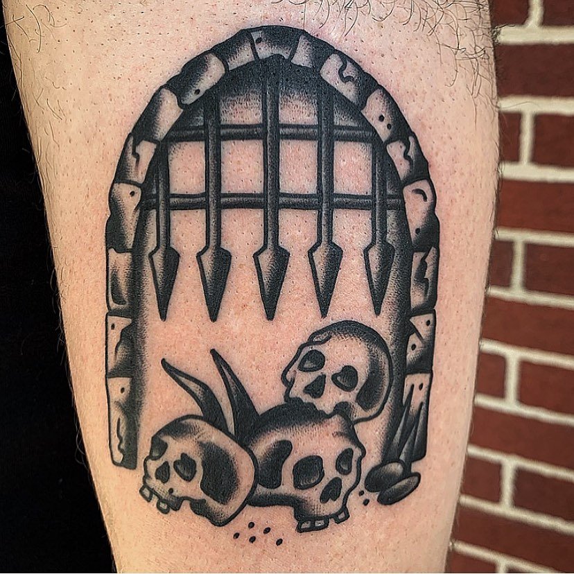 Tattoo of a graveyard scene featuring three skulls, blades of grass, and a fence with pointed pickets.