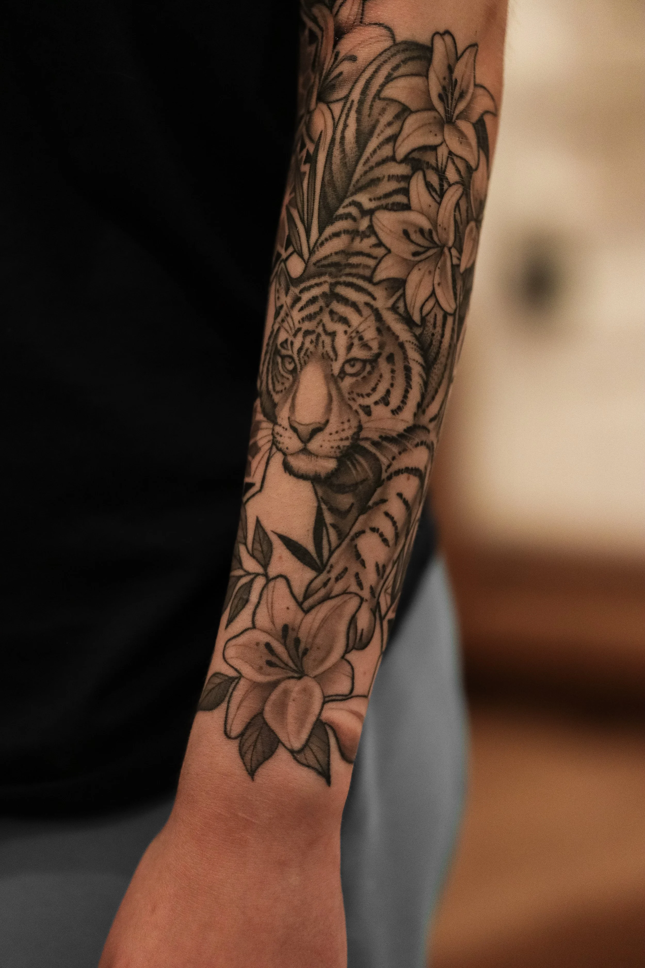 A person's arm featuring a detailed black and gray tattoo of a tiger's face surrounded by flowers and leaves.