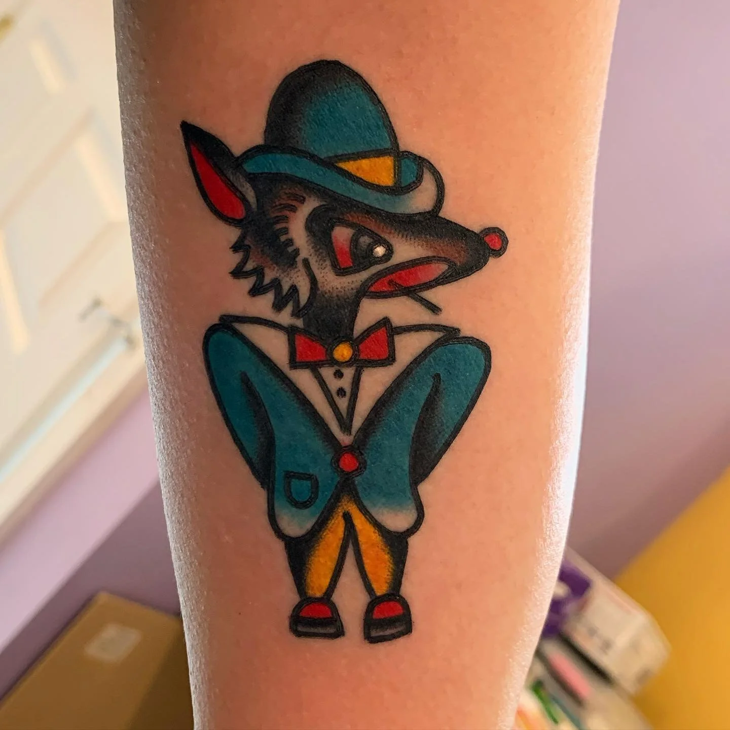Colorful tattoo of a cartoon mouse dressed as a clown, wearing a top hat, bow tie, and clown shoes, with large ears and a long snout.