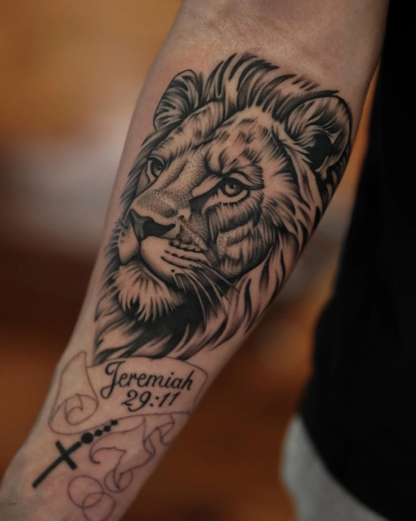Black and gray tattoo of a lion's face on a person's arm, with the name Jeremiah and the date 29-11 written below.