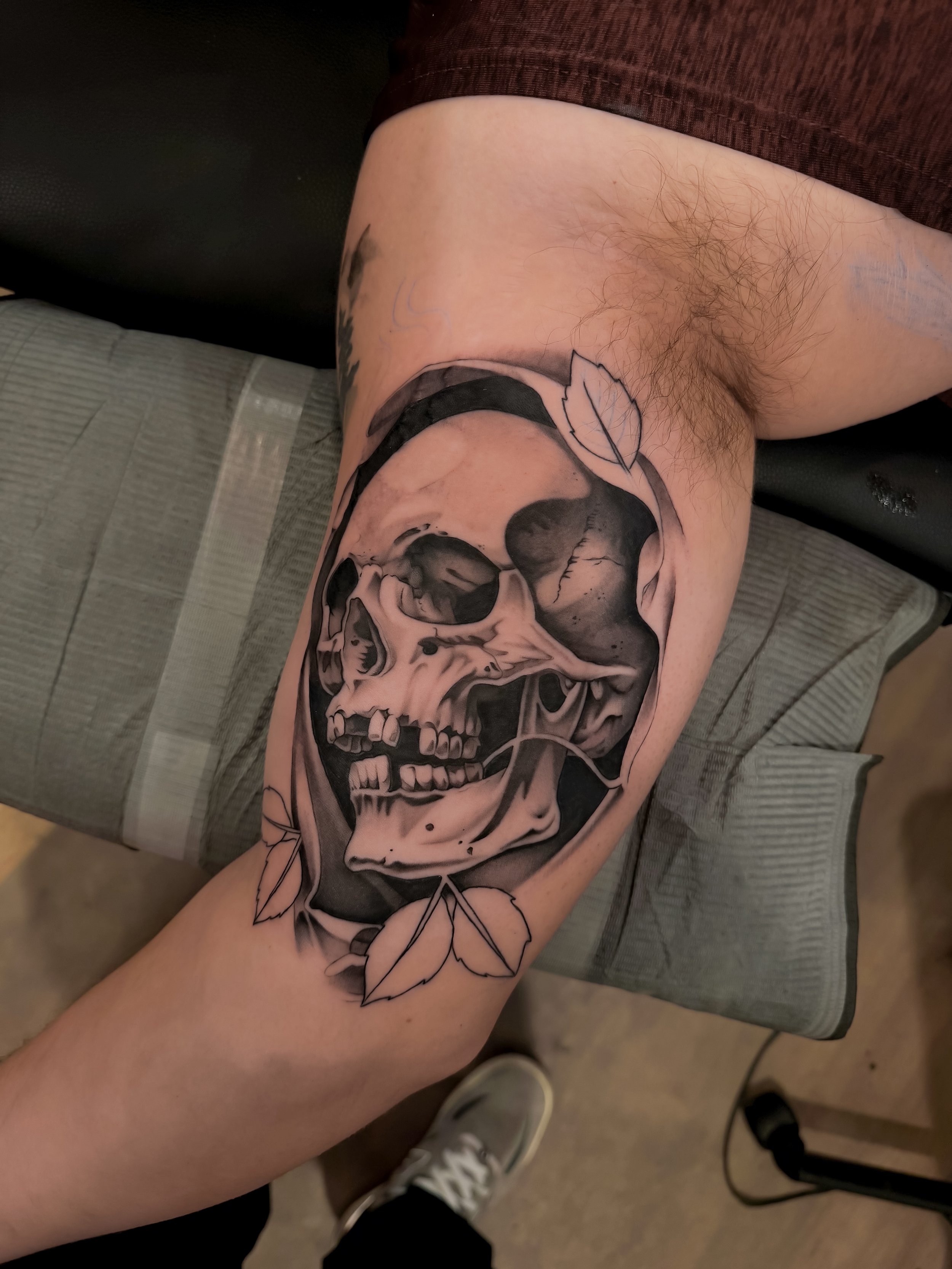 Tattoo of a skull with leaves on a person's thigh.