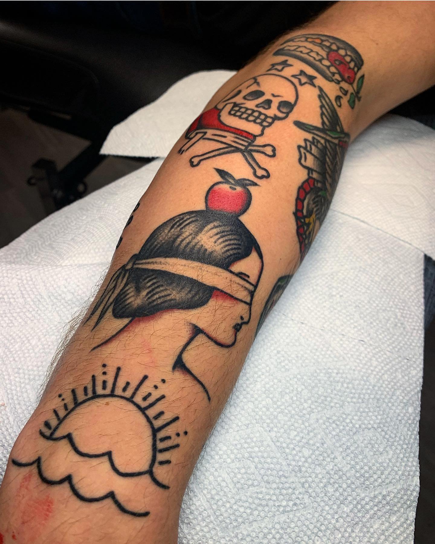Tattoo on forearm featuring a woman's face with closed eyes and dark hair, a sun with radiating lines, an apple with leaf, a skull with stars on the forehead and crossed bones below, a skull wearing a red bandana, a chain and a red apple, and additio