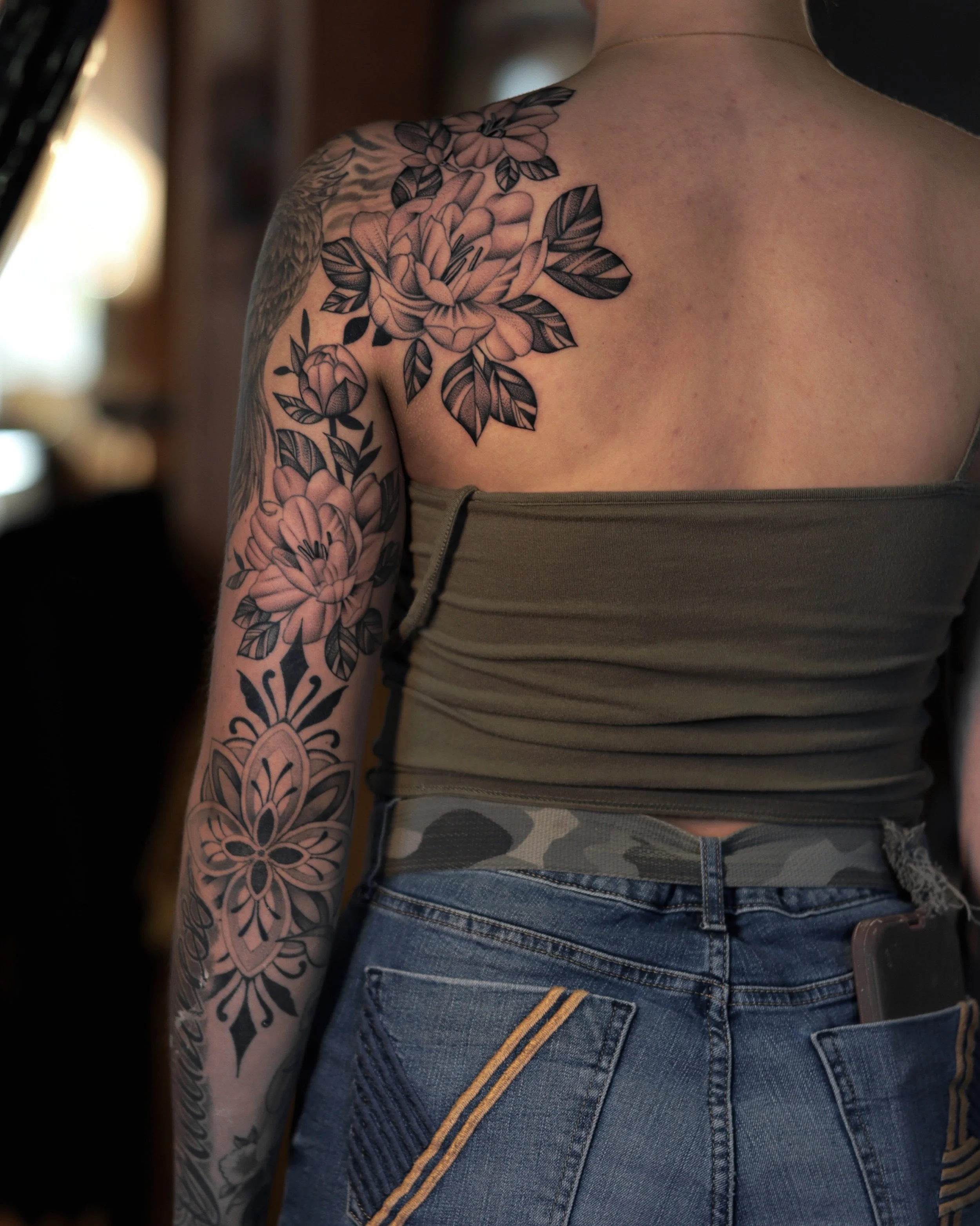 A person with a floral tattoo sleeve on their arm, wearing a tank top and denim shorts, standing in a dimly lit indoor space.
