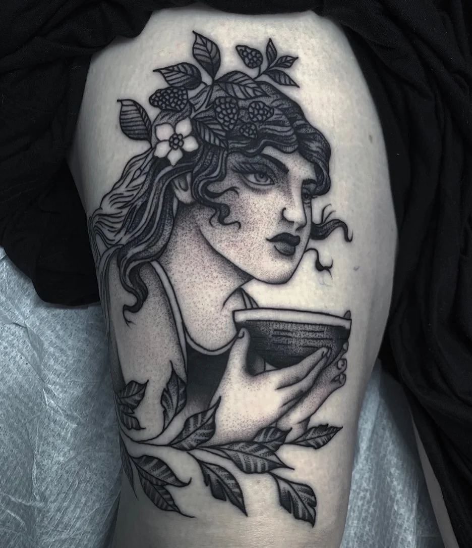 Black and gray tattoo of a woman holding a cup, with flowing hair, leaves, and flowers, on a person's thigh.