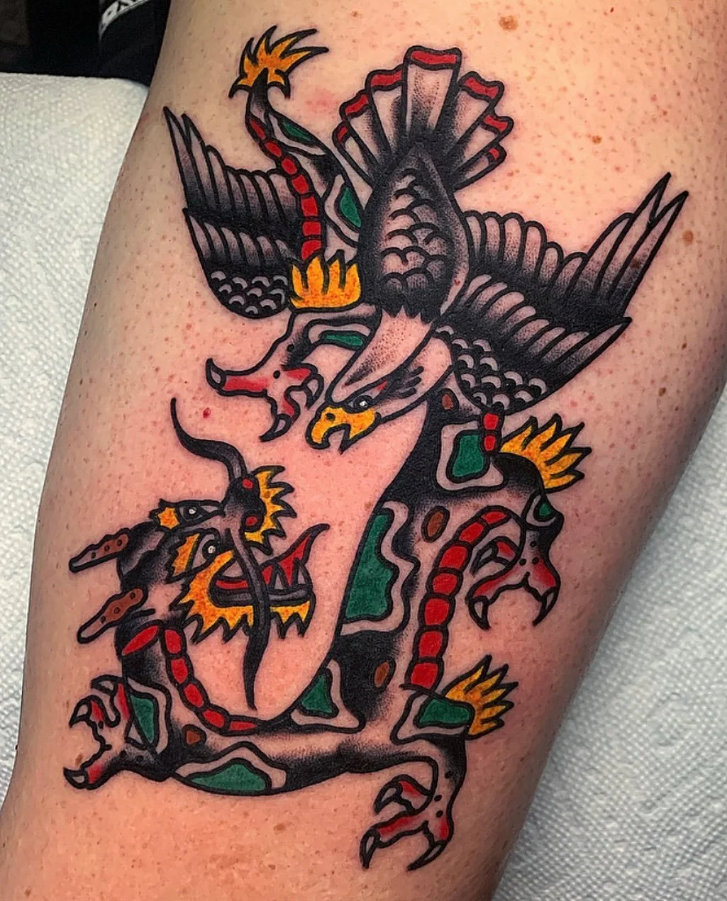 A traditional-style tattoo of a dragon intertwined with a phoenix, featuring bold black outlines, red, yellow, and green details.