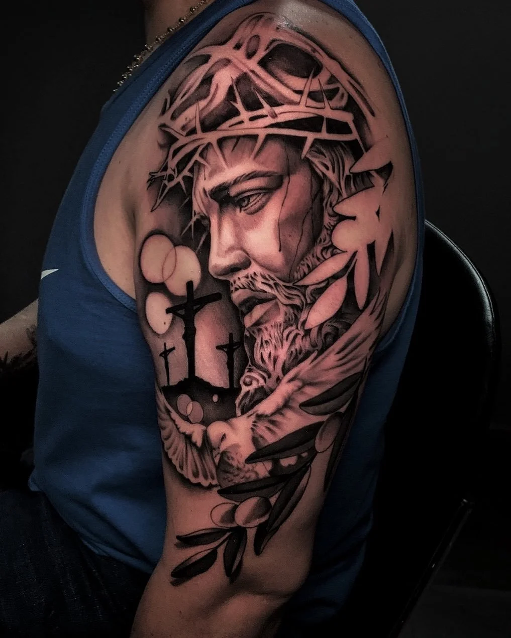 Black and gray tattoo of Jesus Christ wearing a crown of thorns, with a contemplative expression, surrounded by abstract elements including crosses, circles, and leaves.