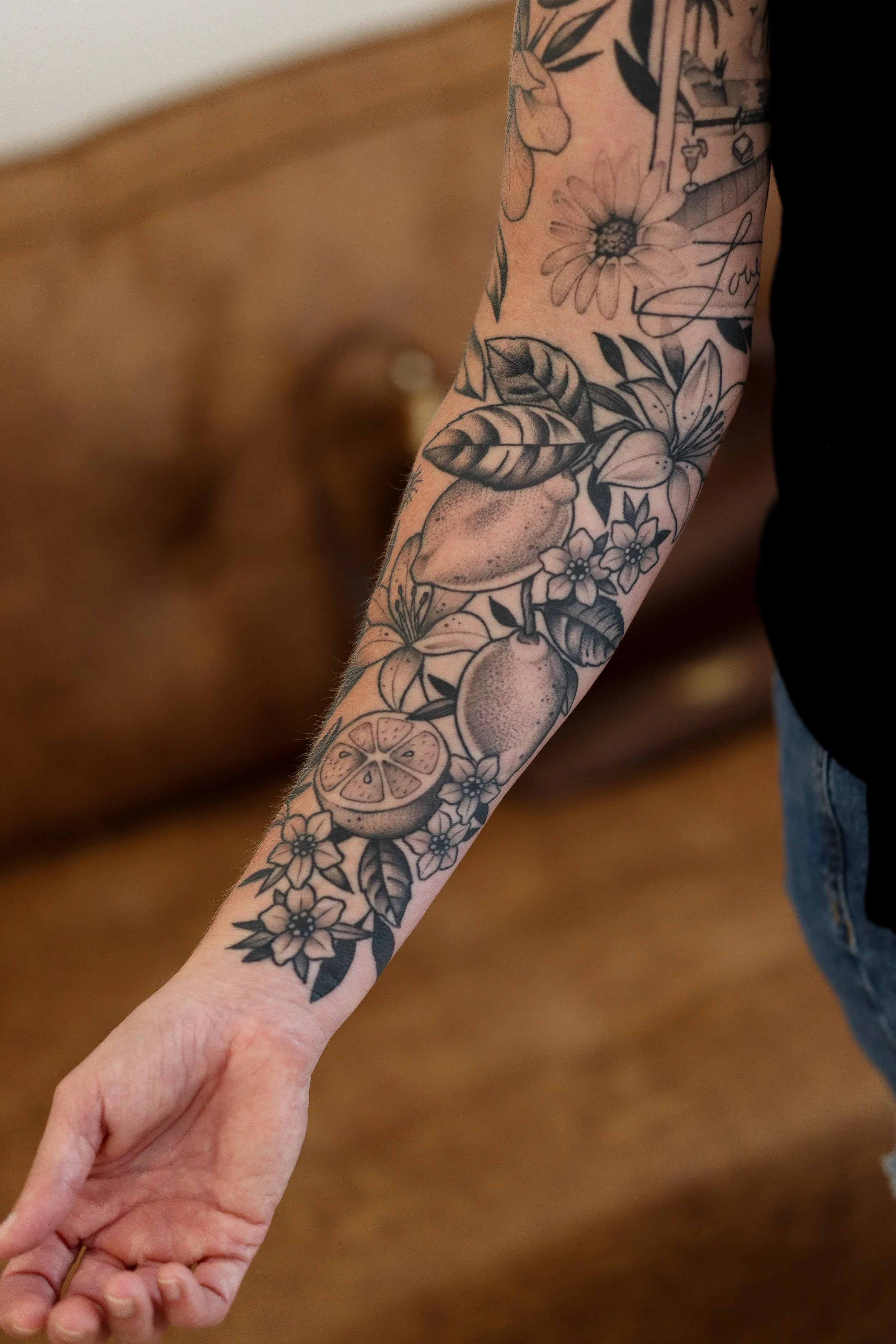 A person's arm with a floral tattoo featuring various flowers, leaves, and a citrus fruit, with some script writing visible on the tattoo.