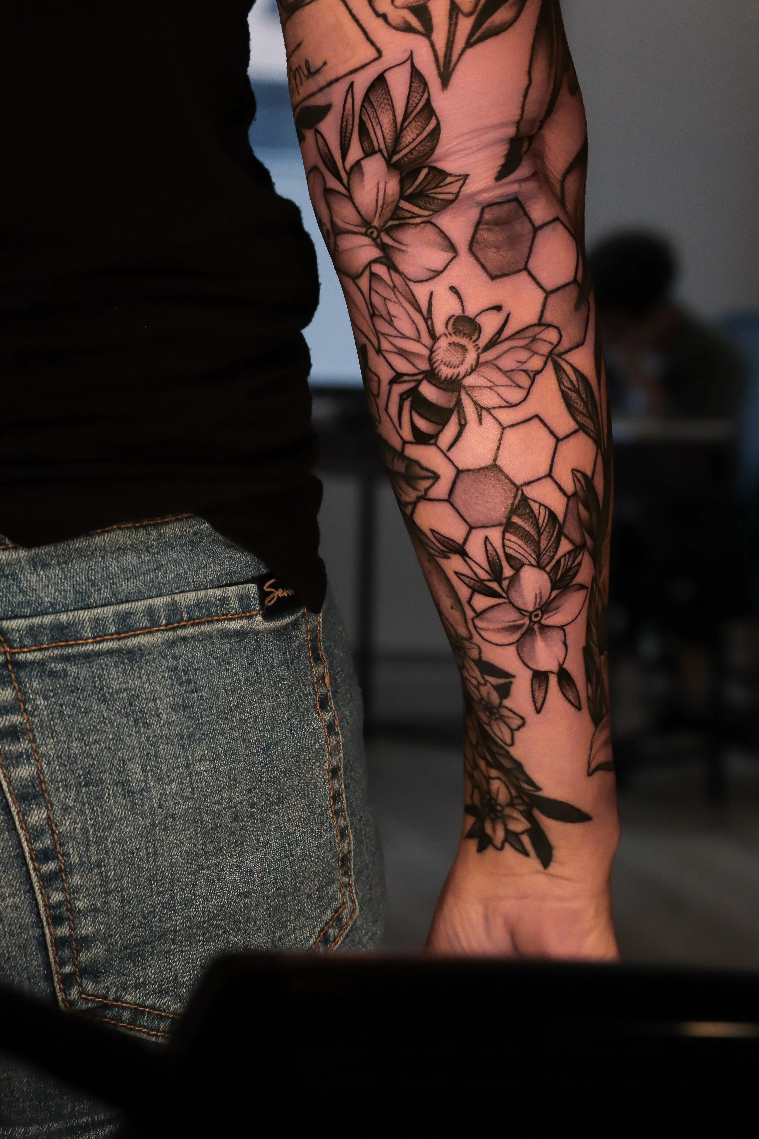 A person's arm with a detailed black and gray floral and bee tattoo, their side partially visible, wearing a black shirt and blue jeans. The background is indoors and slightly blurred.