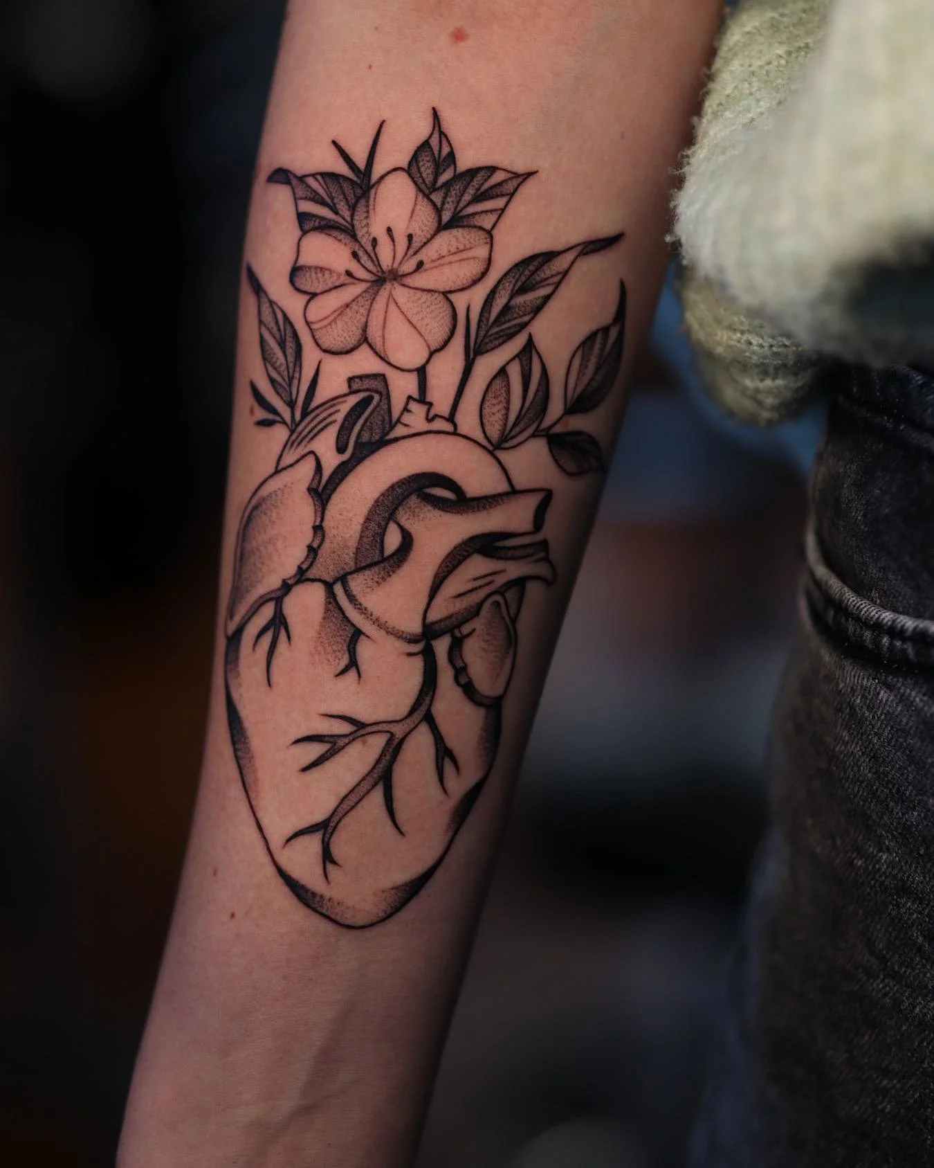 Black and gray tattoo of a heart with a flower and leaves on a person's inner forearm.