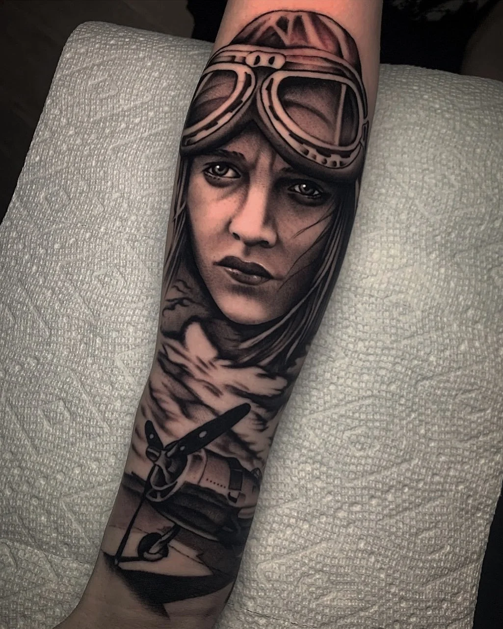 A detailed black and gray tattoo of a woman wearing goggles on her forehead, with a serious expression, above a scenic background with mountains, clouds, and a sickle, on a person's forearm.