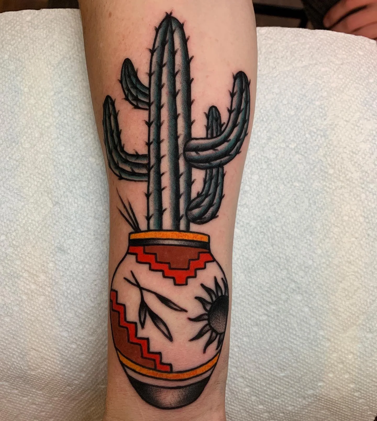 A tattoo of a tall saguaro cactus in a red and orange pot, with a design resembling a Mexican blanket pattern, and a sun with rays below the cactus.