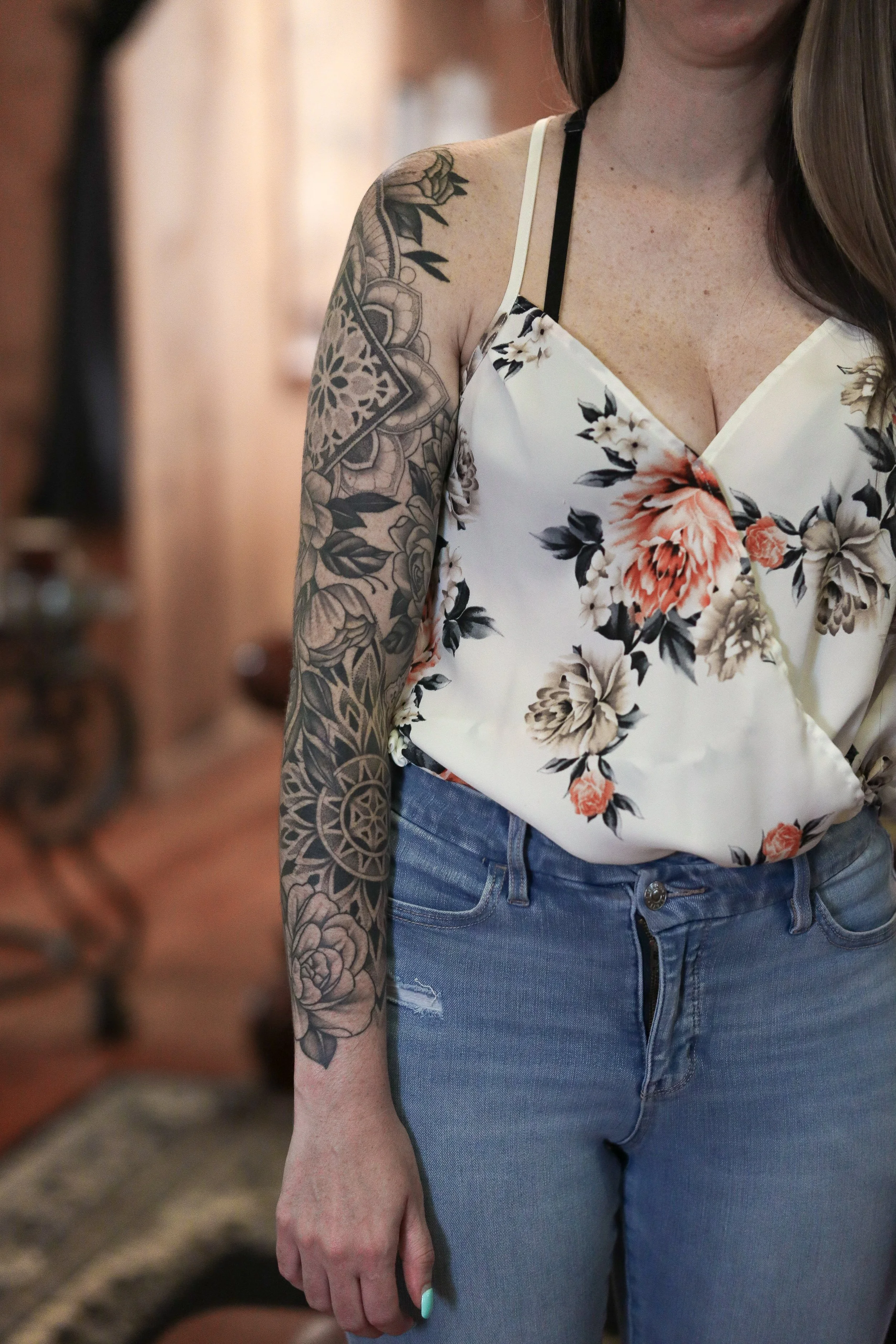 Close-up of a woman wearing a floral sleeveless top and high-waisted blue jeans, showing a tattoo on her upper arm.