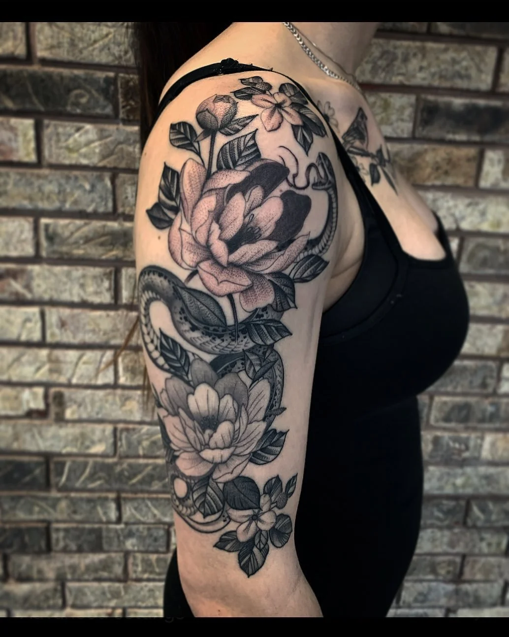 Black and gray tattoo sleeve featuring flowers and a snake on a woman's upper arm, with a brick wall background.