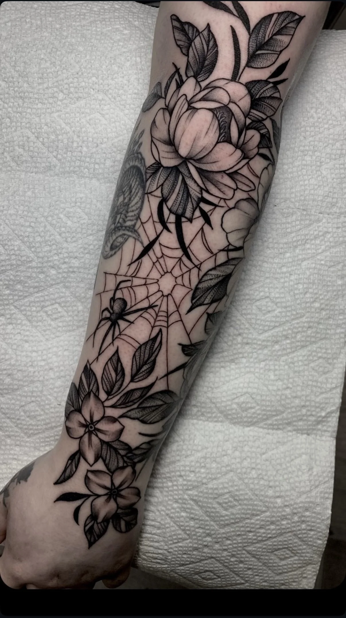 Black and gray floral tattoo featuring a rose, flowers, leaves, a spider web, and a spider on a person's forearm.
