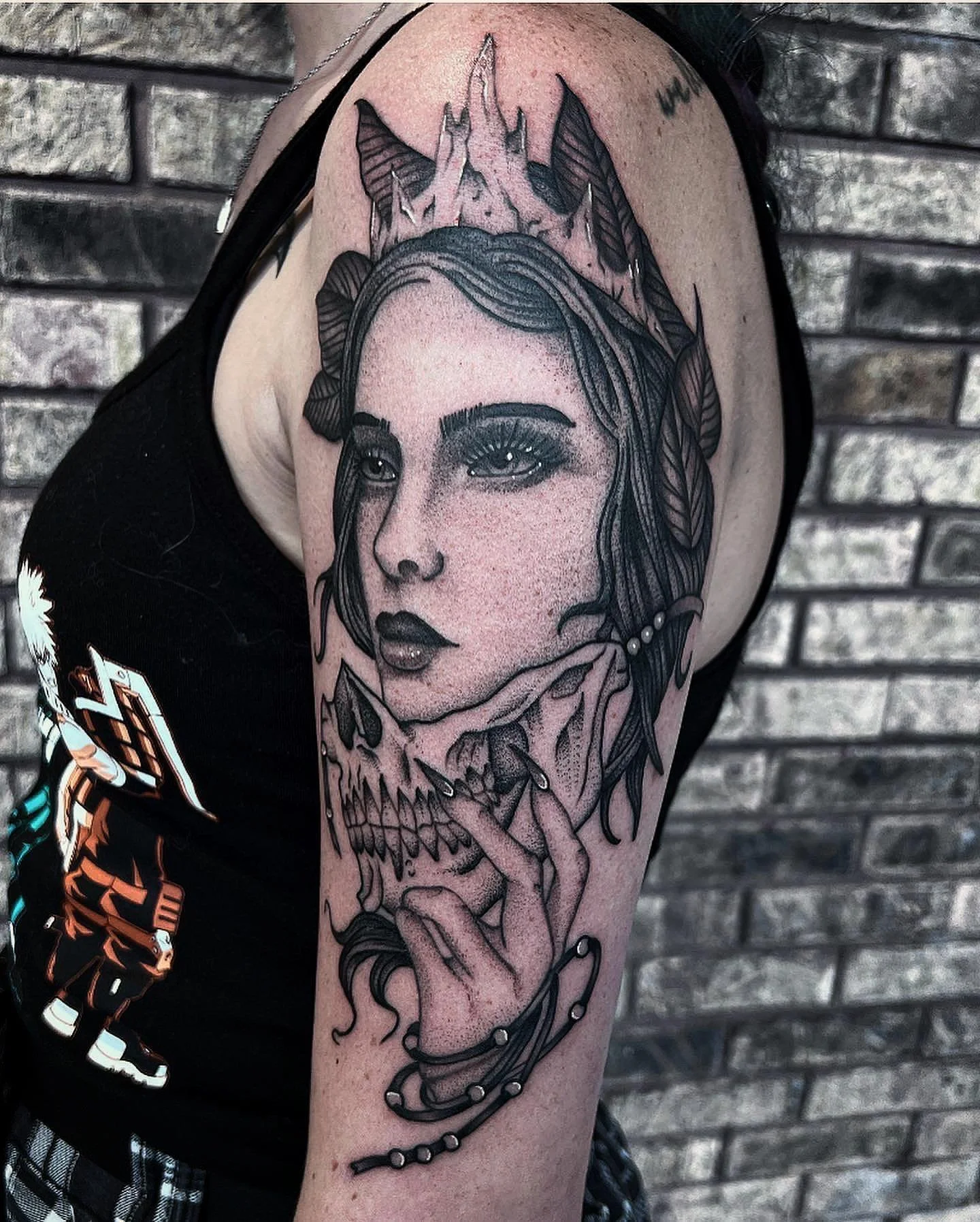 Tattoo of a woman's face with long hair, heavy makeup, and a serene expression, surrounded by leaves, a butterfly, mountain peaks, and a skull, all rendered in black ink on upper arm against a brick wall background.