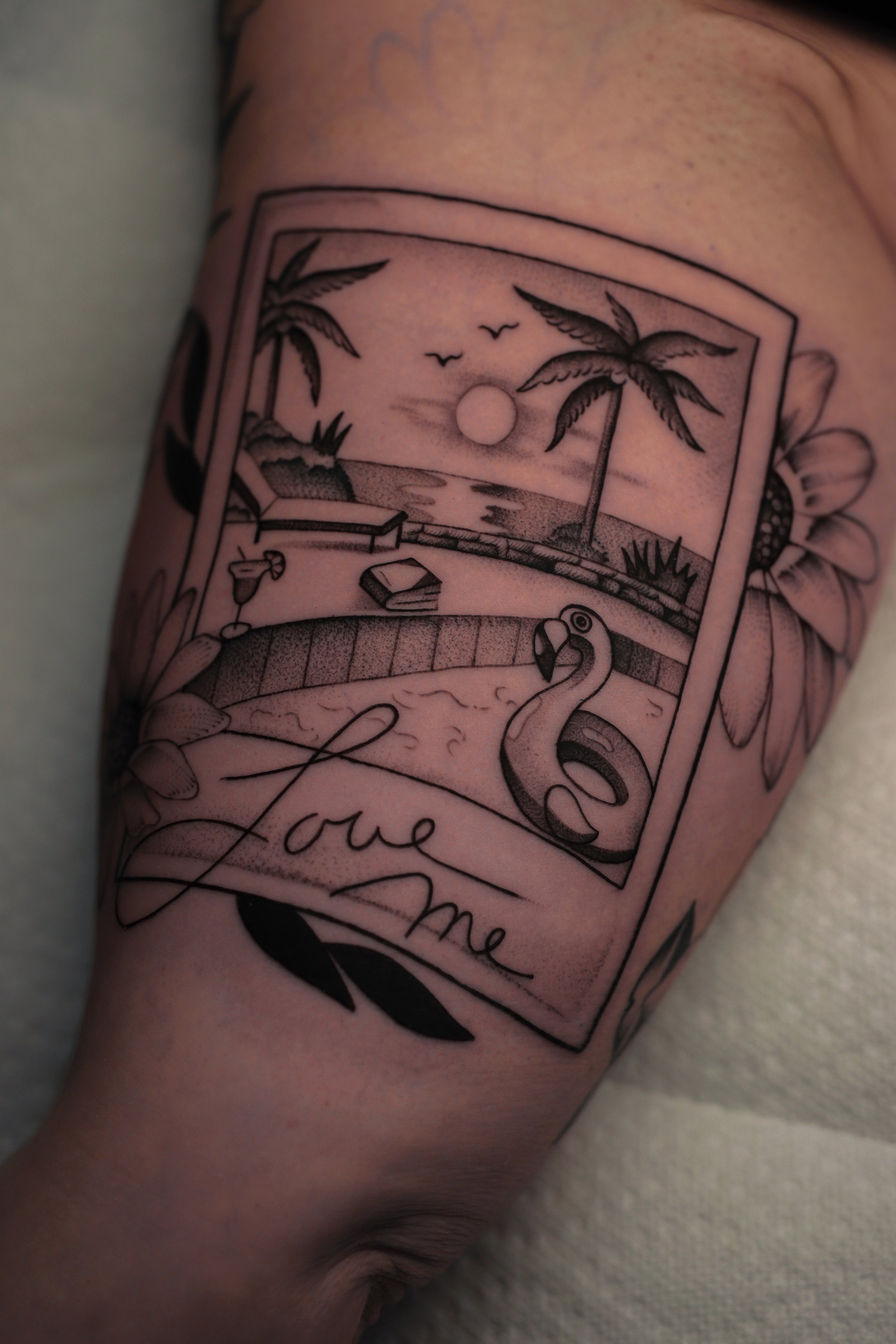 Tattoo of a tropical beach scene with palm trees, a sunset, a swan, and the words 'love me' on a person's shoulder.