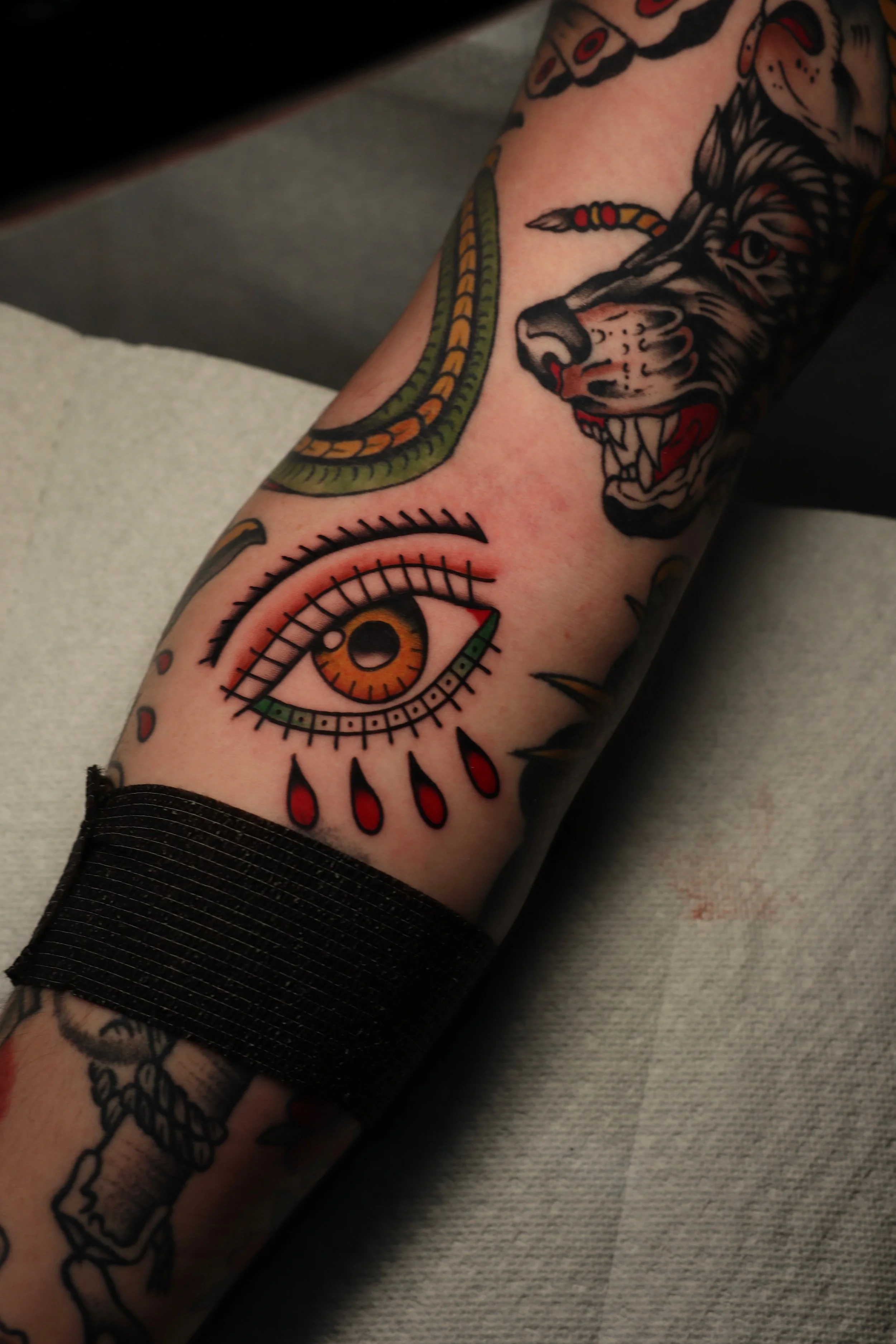 Tattoo of a fierce wolf's head with its mouth open and teeth exposed, surrounded by traditional tattoo elements including an eye with orange and yellow iris, and ribbon-like patterns, on a person's forearm.