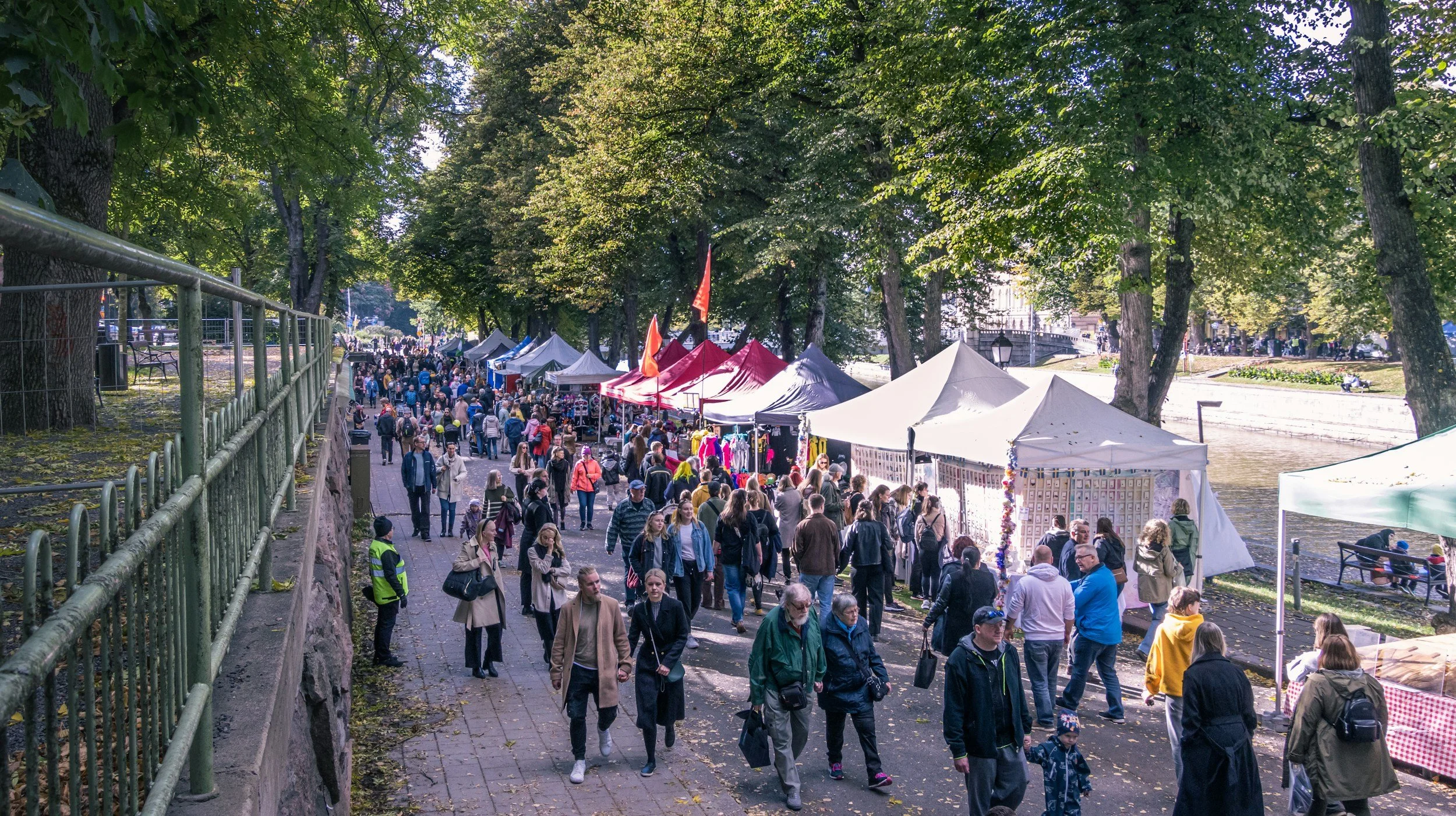 Crowd of people walking along a riverside festival or market with tents, trees, and a lamppost nearby.