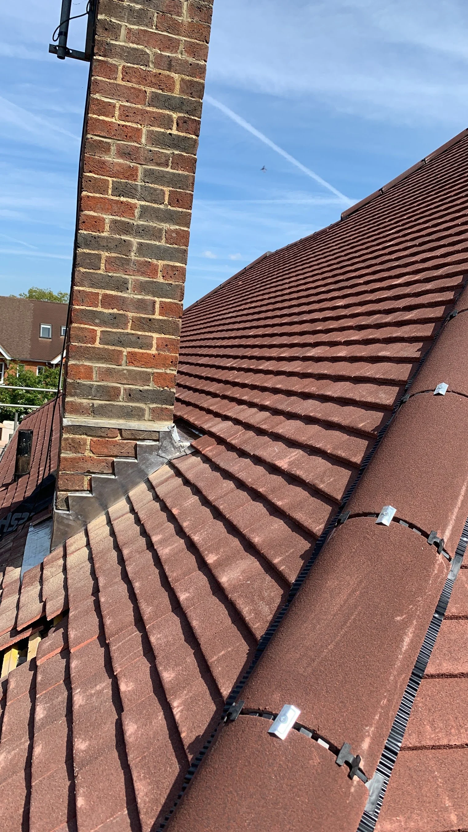 Close-up view of a sloped red roofing with a chimney. The background shows a blue sky with a jet trail and a small airplane.