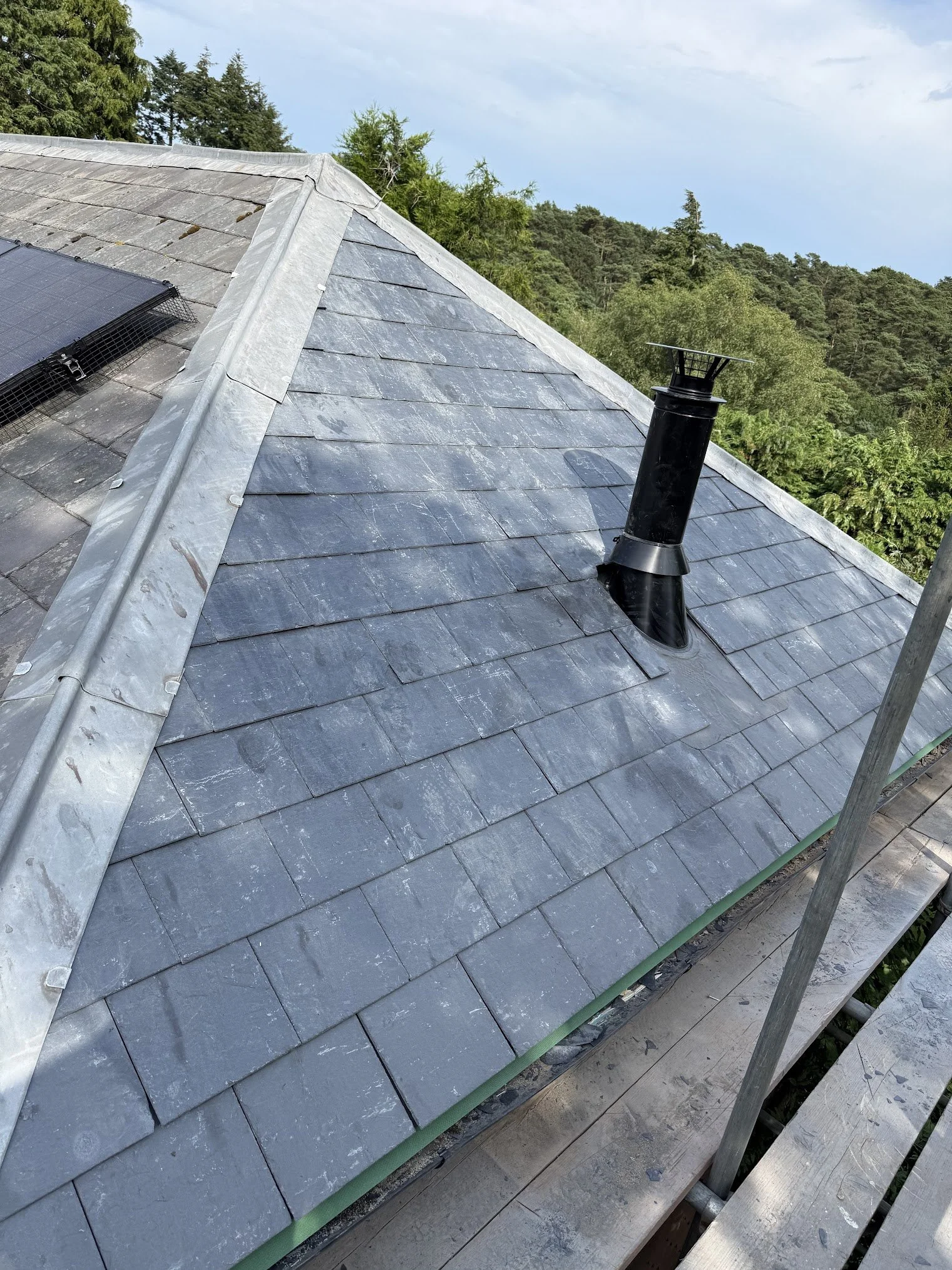 A roof with gray slate tiles, a chimney pipe, and another solar panel on the left side, with trees and a cloudy sky in the background.
