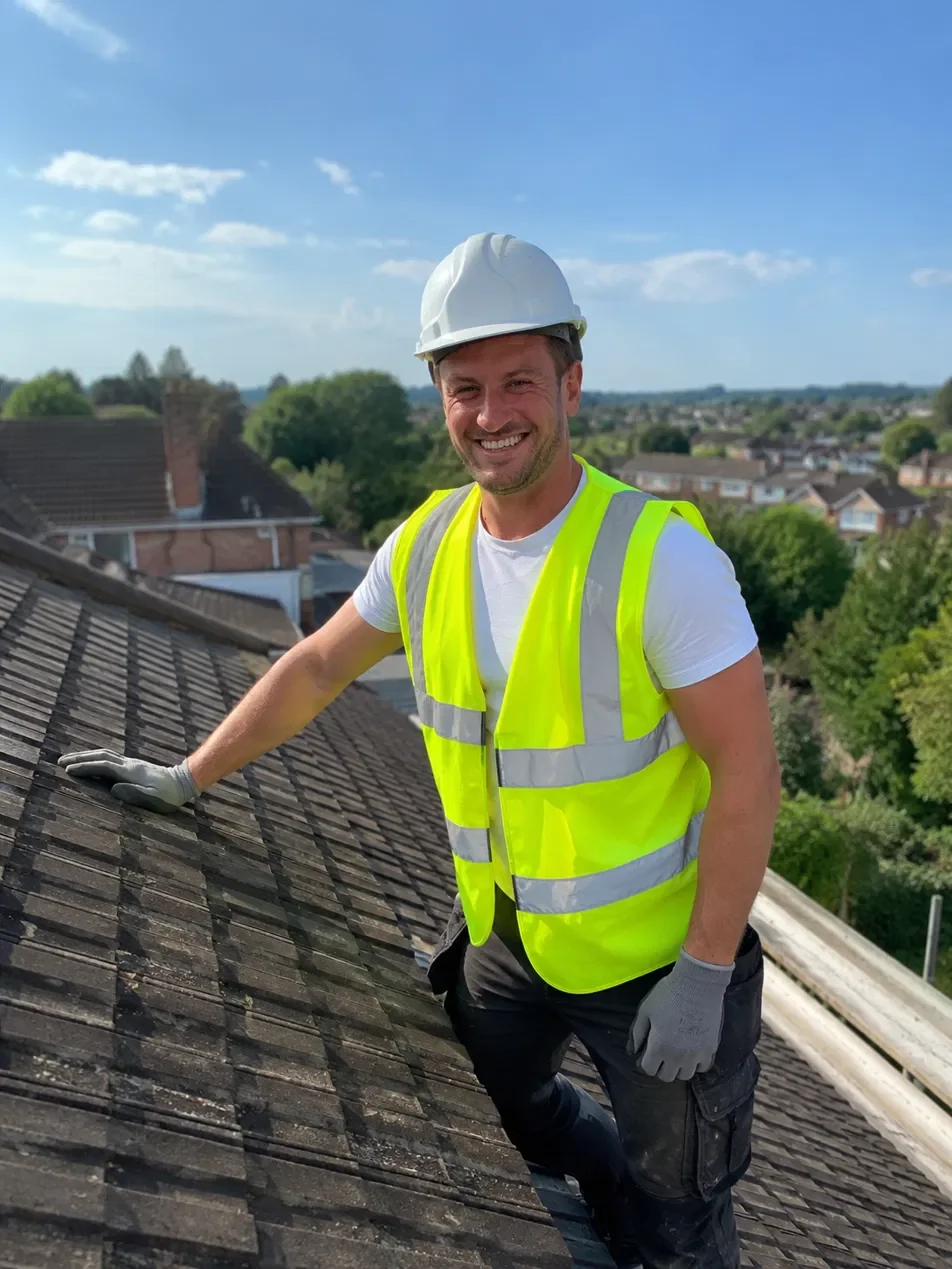 A smiling man in a white hard hat, yellow safety vest, gray gloves, and black work pants, working on a rooftop during the day with a suburban neighborhood and blue sky in the background.